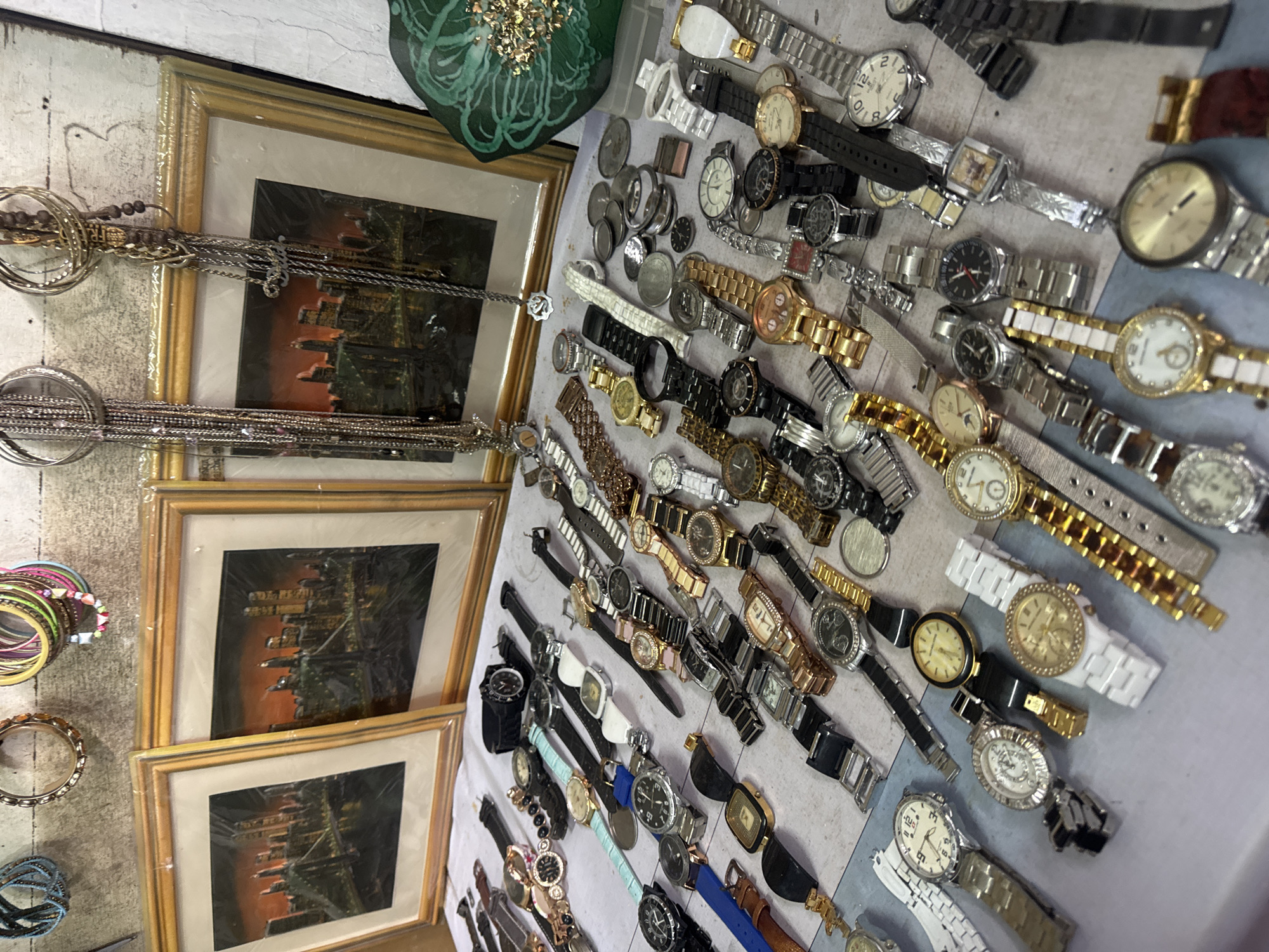 The image shows a close-up, high-angle view of a variety of wristwatches laid out on a table. The watches are a mix of styles and colors, with metal bands and leather straps. Some of the watches have visible faces and hands, while others are less clear. In the background, two framed pictures of a city skyline at dusk are visible, along with some hanging jewelry, including bracelets and necklaces. There is also a green, decorative item in the far right background. The overall impression is of a market stall or a display of goods for sale. The scene does not provide enough information to determine a specific location within Denpasar, Indonesia.
