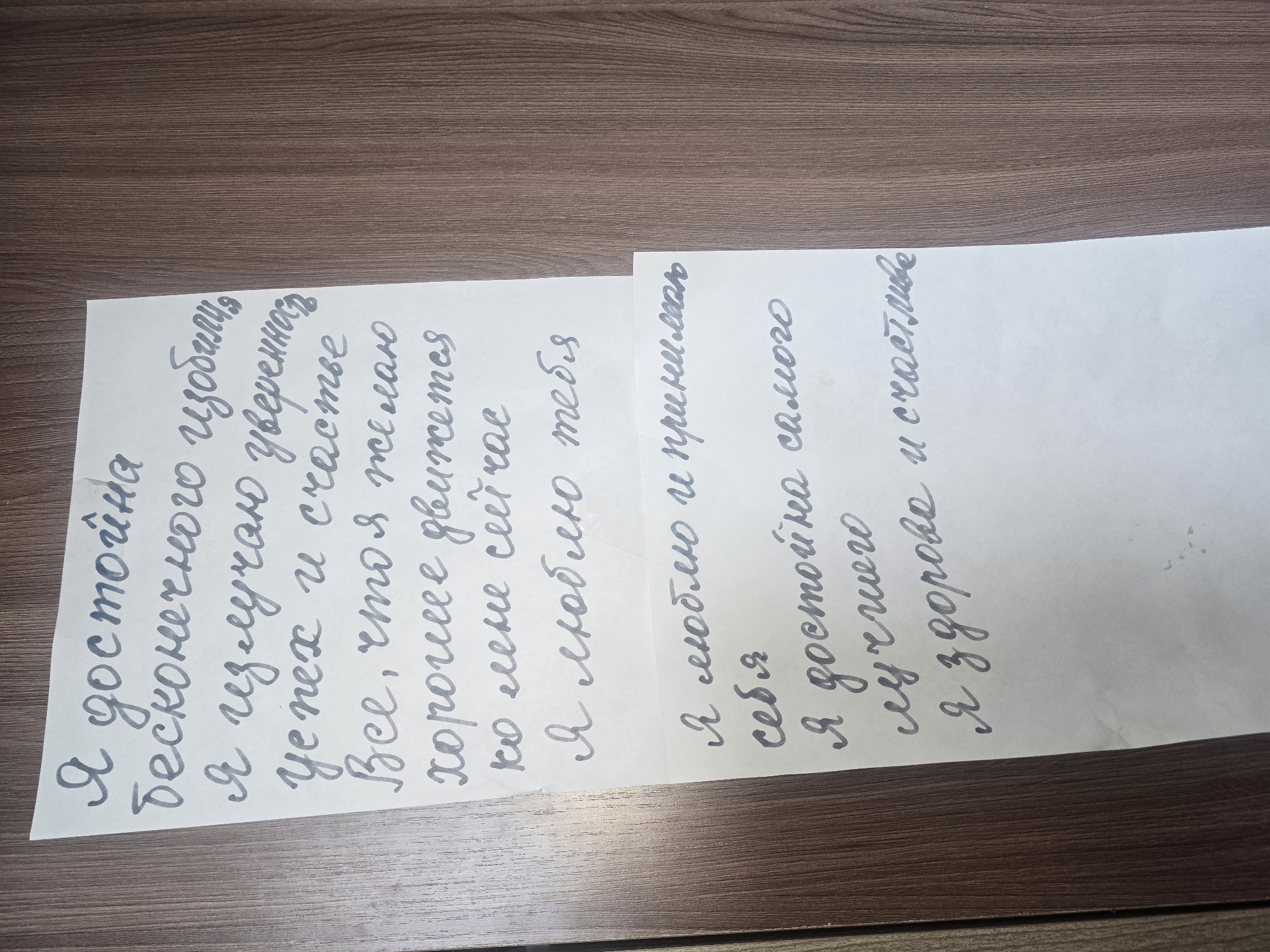 The image shows a piece of paper with handwritten Russian text on a wooden background. The text is written in grey ink. The paper is positioned vertically on the left side of the frame. The wooden surface has a dark brown color with vertical grain patterns. The text appears to be a personal affirmation or reflection. The handwriting is cursive and somewhat stylized. The top of the paper has a slight bend. The lighting suggests an indoor environment. The specific location in Ulan-Ude, Russia cannot be determined from the provided image.