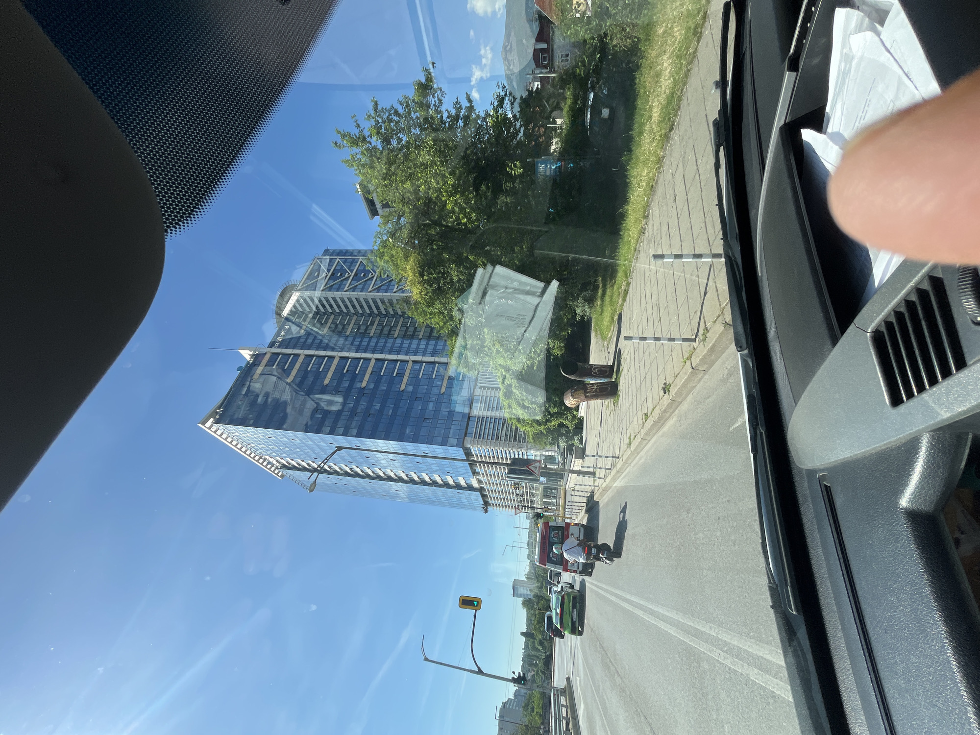 A clear, sunny day is depicted from the perspective of someone inside a vehicle, looking forward through the windshield. The scene captures a street in a city with a prominent, modern skyscraper featuring a glass facade dominating the right side of the frame. Traffic is visible on the road, including a motorcycle with a rider, a green car, and other vehicles further ahead. A traffic light with a green signal is visible on a pole to the left. The sidewalk along the road has a paved surface with bollards. Trees and other buildings are also present in the background, and mountains can be seen in the far distance under a bright blue sky with wispy clouds. The dashboard of the vehicle is partially visible in the foreground.