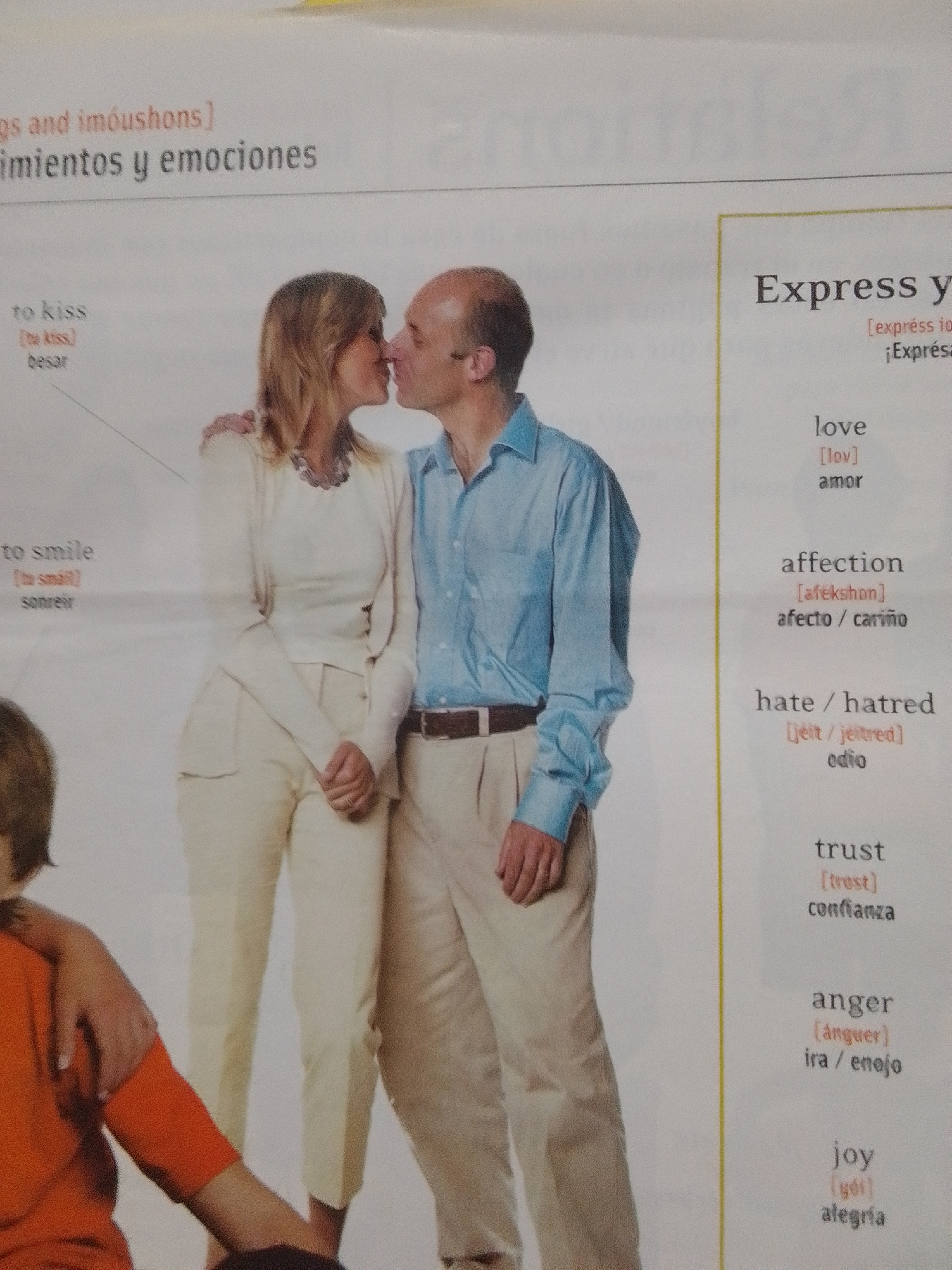 The image shows a book page with text and a photograph. The photograph depicts a man and a woman kissing. The man is wearing a blue collared shirt and tan pants, while the woman is wearing a cream-colored sweater and pants. To the left of the photograph, text lists "to kiss" with phonetic spelling and Spanish translation. To the right, a section titled "Express y" lists words like "love," "affection," "hate/hatred," "trust," "anger," and "joy" with phonetic spellings and Spanish translations. The background of the page is white and yellow. There is no information or imagery related to the city of La Tagua, Venezuela in this image.