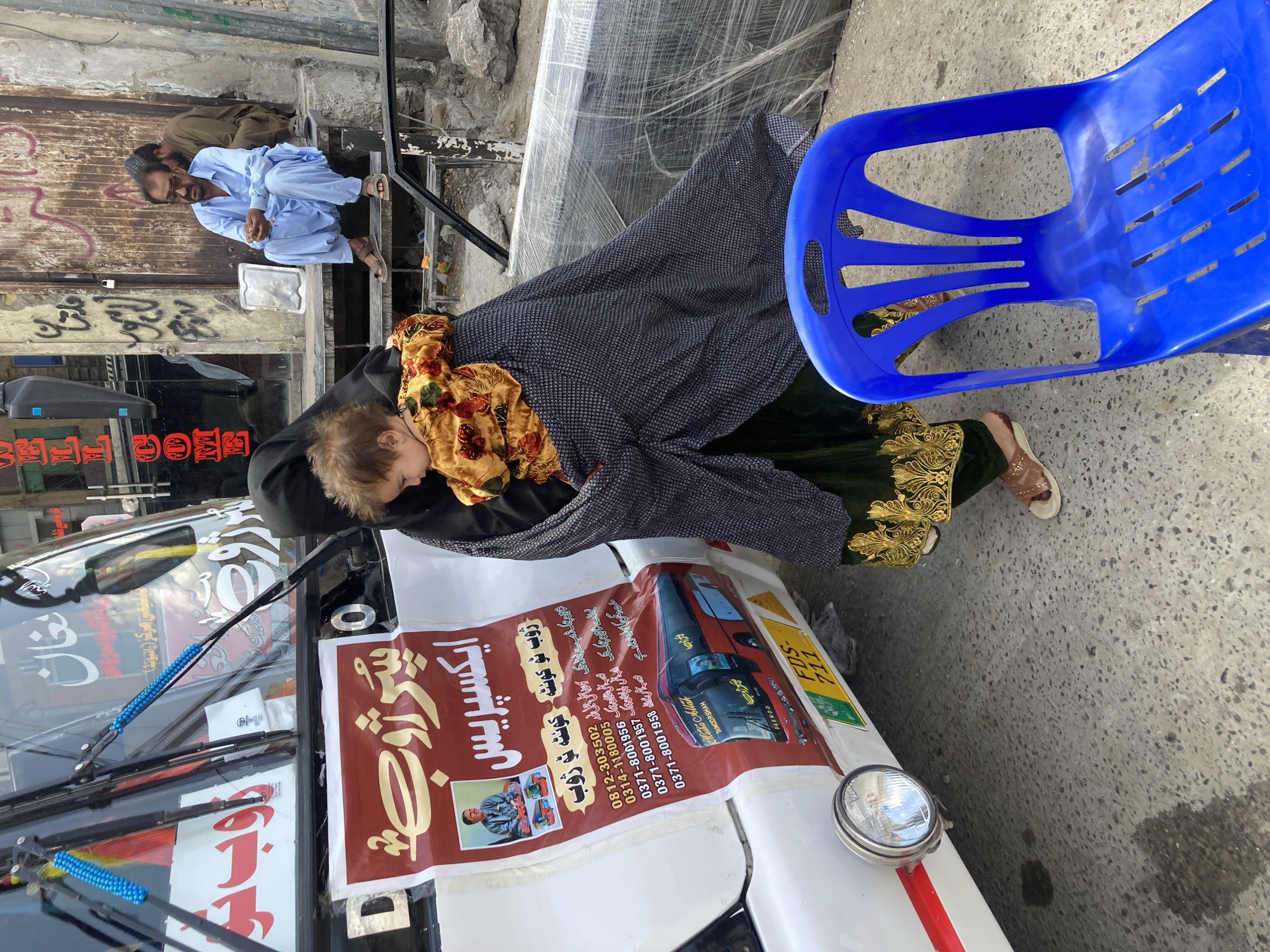 The image captures an outdoor urban street scene in Quetta City Tehsil, Pakistan, under bright daylight. In the foreground, a woman, dressed in a dark, abaya-like garment, cradles a baby or toddler wrapped in a colorful, floral-patterned blanket. The woman's feet are visible, wearing light-colored sandals. To the right of the woman and child is a bright blue plastic chair.
Behind them, a white vehicle, likely a bus or large van, dominates the left side of the frame. A large, reddish-brown banner advertising "ایکسپریس" (Express) transportation services is prominently displayed on the front of the vehicle. The banner explicitly states routes like "ژوب ٹو کوئٹہ کوئٹہ ٹو ژوب" (Zhob to Quetta, Quetta to Zhob) and includes several phone numbers (e.g., 0812-303502, 0371-8001956), names such as "Matha Allak" and "TAIMOOR SHAH," and the brand "DAEWOO." A vehicle registration plate, "FDS-711," is also visible near a headlight. The windshield of the vehicle has other stickers and decorations, including blue beaded adornments on the windshield wipers, and the words "WELL COM" are partially visible on a sign.
In the mid-ground, slightly elevated on concrete steps, two men are seated. One man, in a light blue shalwar kameez and prayer cap, faces the viewer. Another man, in a brown outfit, sits behind him. The background features worn building facades with graffiti, hinting at an active street environment. The ground is paved, and there are no clear indications of extreme weather; the scene appears calm and part of daily life. The overall impression is one of a busy, lived-in streetscape.