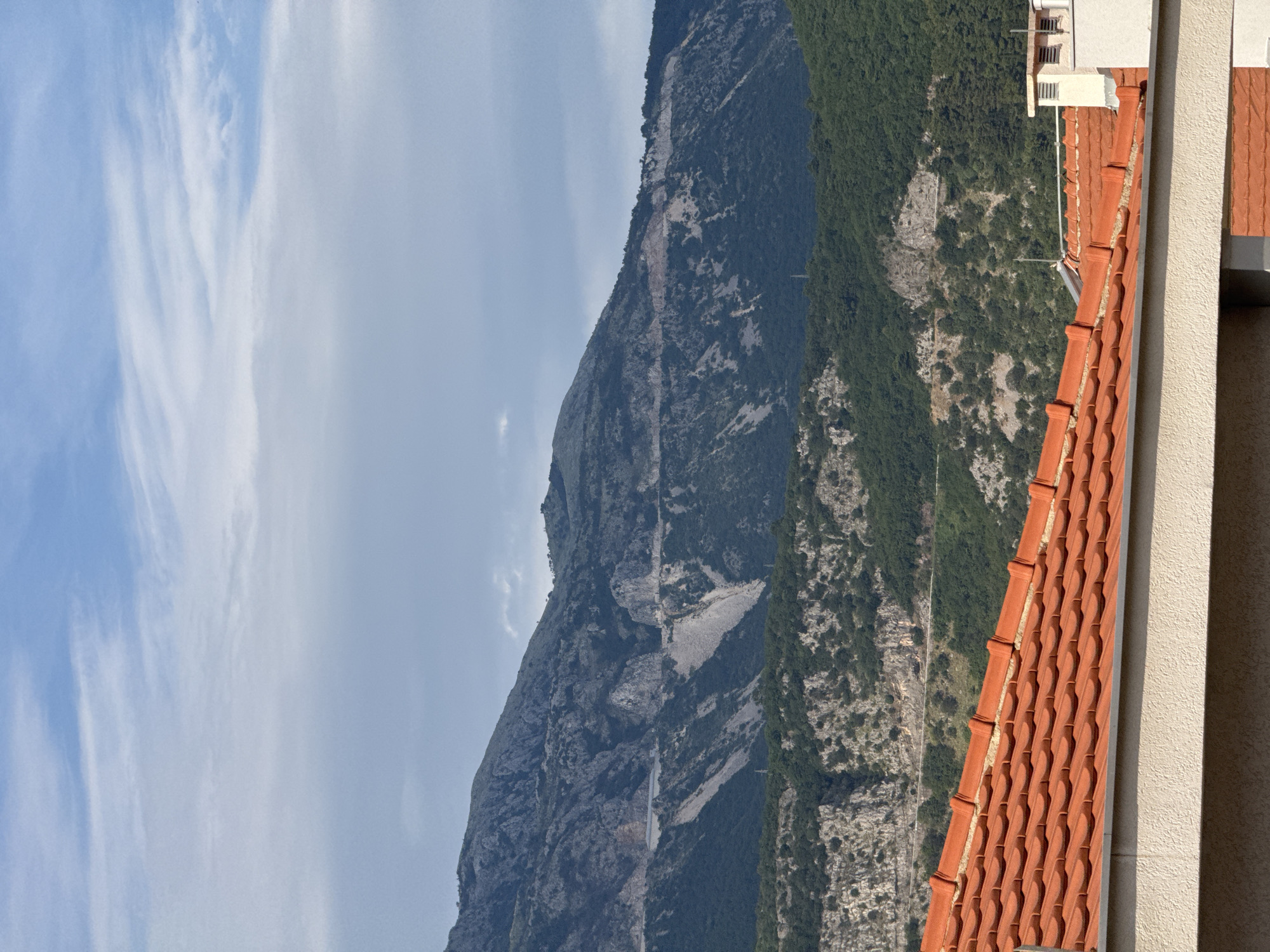 The image captures a view of mountains from a rooftop in Budva, Montenegro. In the foreground, a terracotta tiled roof dominates the lower part of the frame. The tiles are arranged in overlapping rows, showing a light texture and a warm orange hue. A white soffit runs along the edge of the roof. Behind the roof, the landscape rises in stages. The middle ground features a lush green mountainside dotted with trees and interspersed with rocky, barren patches. A winding road is visible carved into the side of a more distant mountain. The background is dominated by a large, imposing mountain range with rugged, rocky slopes and sparse vegetation. The sky above is a mix of pale blue and wispy white clouds, suggesting a partly cloudy day. A white chimney with ventilation pipes is visible on the right side of the roof.