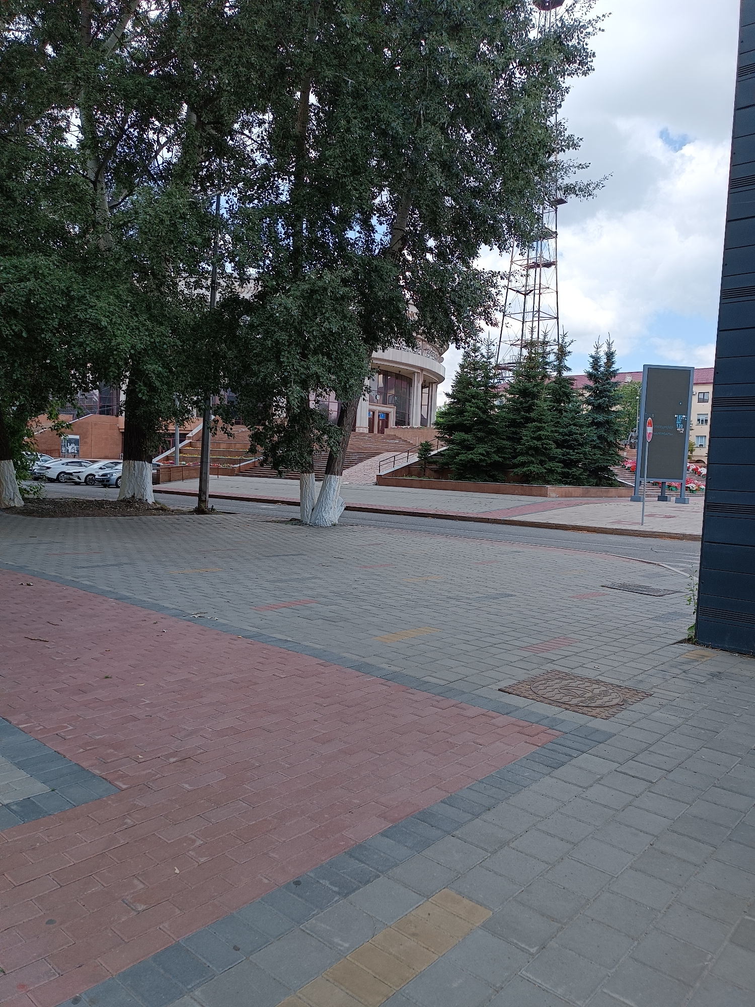 In Qarağandı city administration, Kazakhstan, the image captures a street scene with a paved sidewalk and a road. The sidewalk is constructed with interlocking pavers in varying colors, including red, gray, and yellow. Trees with white-painted trunks line the edge of the sidewalk, separating it from the road. In the background, a large building with a curved facade and grand entrance stairs is visible. Further in the background, a tall lattice tower stands against a cloudy sky. Several evergreen trees are planted in front of the building. Cars are parked on the side of the road. The scene appears to be a public space, possibly a plaza or a street in front of a municipal building, on an overcast day.