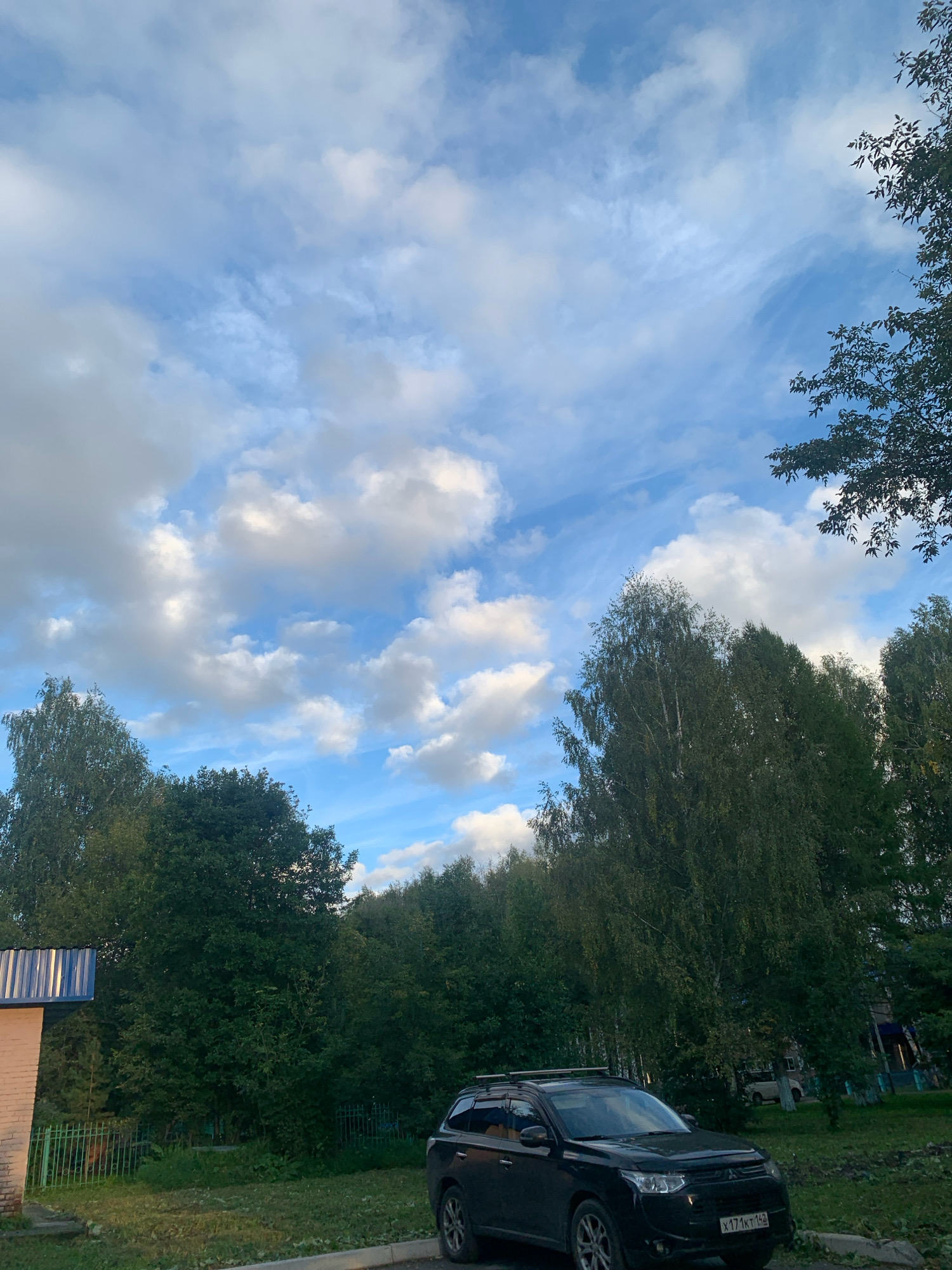 In Prokopyevsk, Russia, this image captures a serene outdoor scene under a partially cloudy sky. A black SUV is parked in the foreground on a paved surface, its license plate clearly visible. Behind the vehicle, a dense line of green trees stretches across the landscape, suggesting a park or a heavily wooded residential area. To the left, a section of a light-colored brick building with a blue-trimmed roof is visible, indicating a structure within the vicinity. The sky is a mix of blue and white clouds, creating a dynamic backdrop. The overall atmosphere is calm and uneventful, with no people present and no apparent activity.