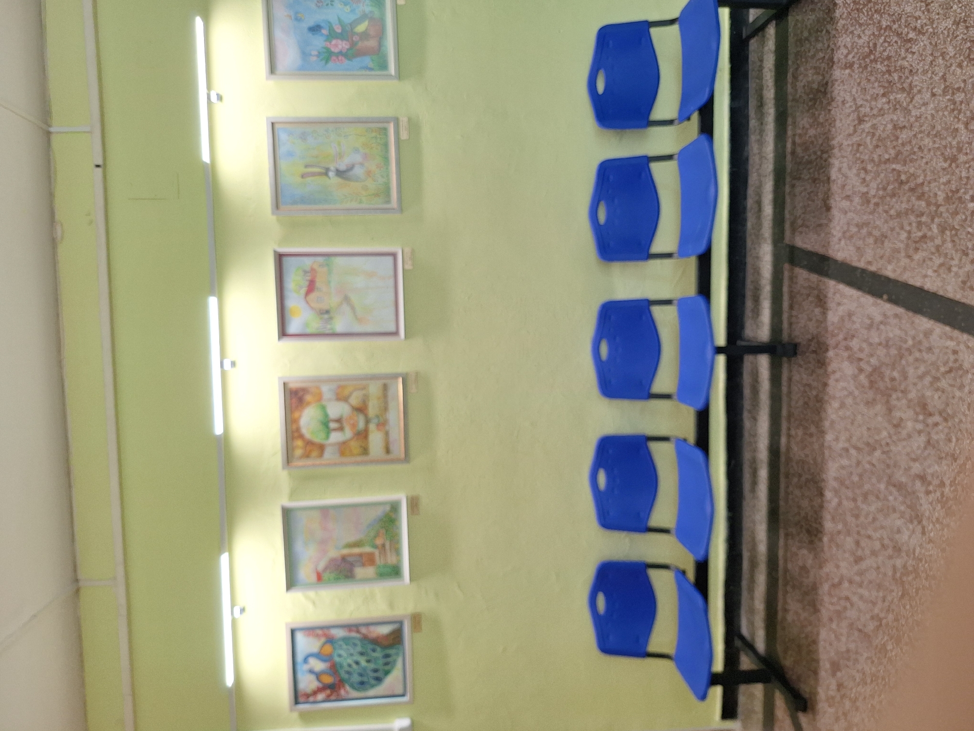 The image shows a wall with several framed paintings and a bench with blue plastic chairs in front of it. The paintings appear to be artwork, with subjects including a peacock, a landscape with a house, a tree, and a rabbit. The wall is painted a pale green, and fluorescent lights are mounted above the paintings. The chairs are simple, molded plastic seats with black metal frames, arranged in a row of five. The floor is a speckled tile. The overall scene suggests a waiting area or a small gallery space. The location is identified as Varna, Bulgaria.