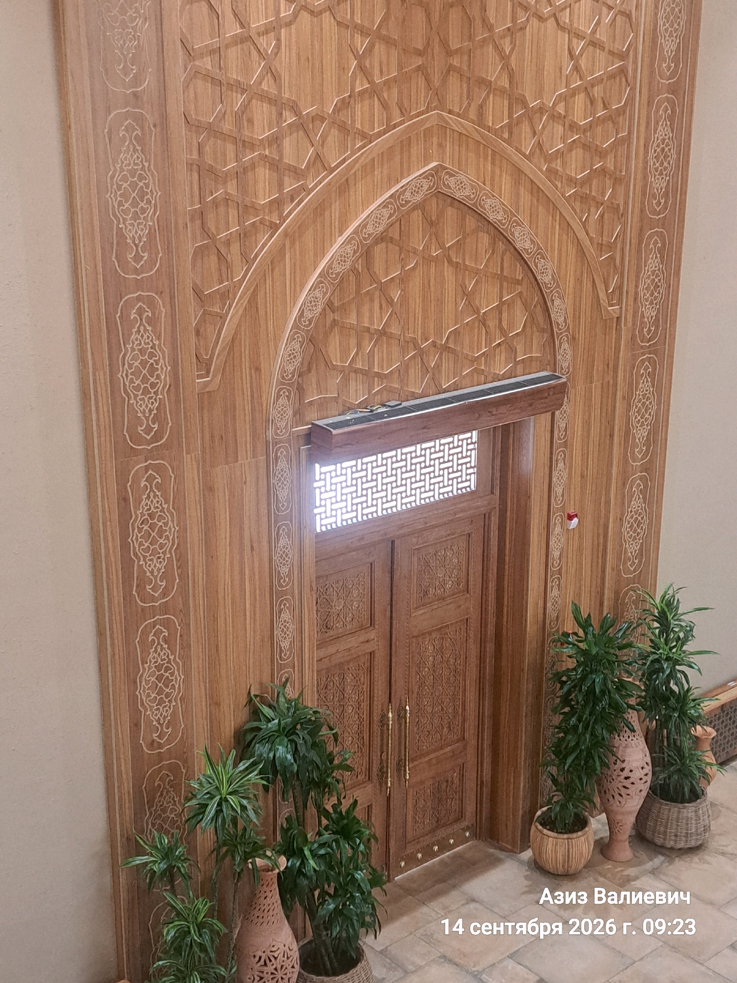 This is a high-angle, medium shot of a wooden doorway with intricate carvings in Tashkent, Uzbekistan. The doorway is framed by an ornate wooden arch and decorated with geometric patterns and decorative inlays. Two tall, green potted plants are positioned to the left of the doorway, and a smaller potted plant is to the right. A decorative clay pot with an intricate pattern is visible in the foreground, to the left of the doorway. The floor appears to be tiled. A timestamp and name are overlaid on the bottom right of the image: "Азиз Валиевич 14 сентября 2026 г. 09:23". There are no people visible in the image. The scene appears to be an interior shot, possibly within a cultural building or a richly decorated hallway.