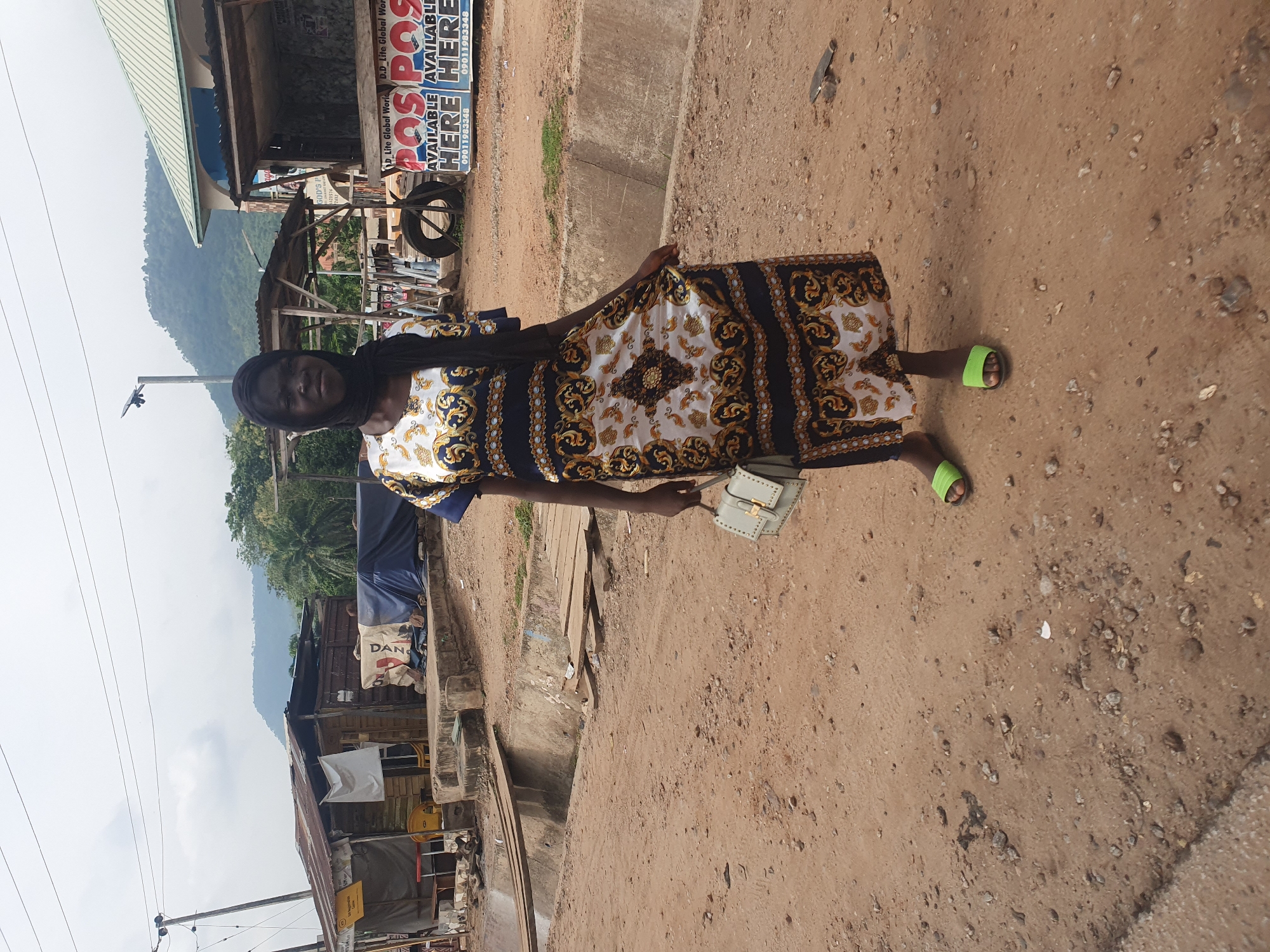 This image depicts a person standing on a dirt road in what appears to be a market or roadside stall area. The person is wearing a patterned dress with black and gold designs and bright green slides on their feet. They are holding a small light-colored purse.
In the background, there are structures that resemble stalls or small shops with corrugated metal roofs. Some are open, revealing goods or materials inside. A prominent sign with the text "POS POS AVAILABLE HERE" and a phone number is visible on one of the structures. There are also some draped tarps and what looks like a tire leaning against a building.
The setting is outdoors, with a dusty, dirt ground covered in small stones and debris. The terrain slopes slightly downward away from the viewer. In the far background, a lush, green mountainside is visible under a hazy, overcast sky, suggesting it might be a cloudy day or a humid environment. The overall impression is a busy, possibly rural or semi-urban, commercial area. The image appears to be taken during the daytime.