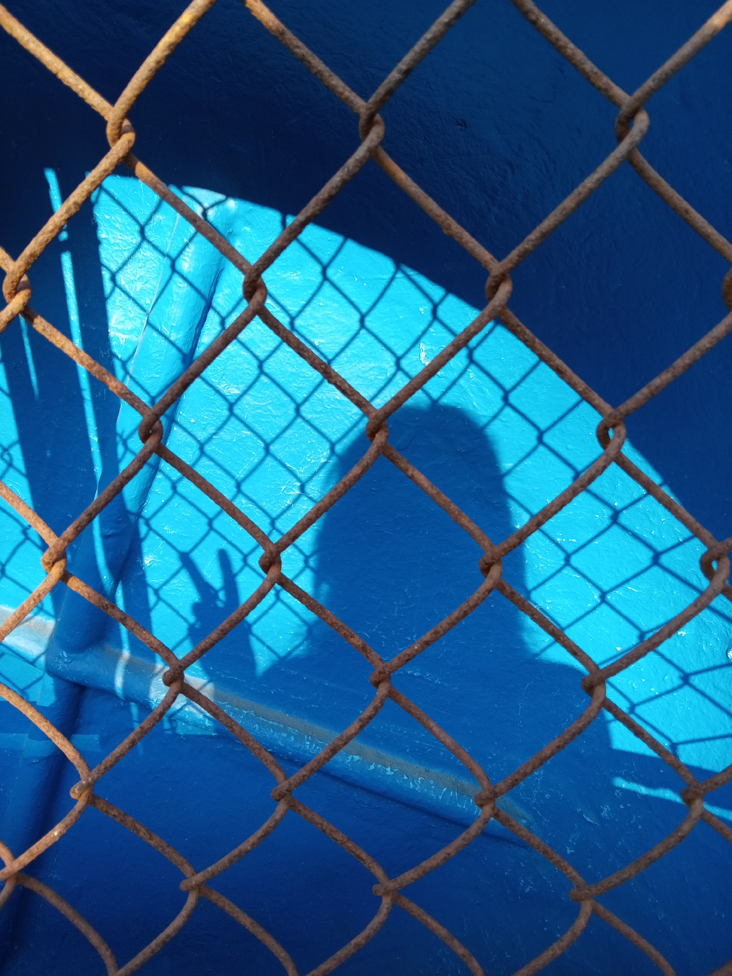 A close-up shot shows a rusty chain-link fence. The fence is positioned in front of a bright blue painted surface. A shadow of a figure making a peace sign with their fingers is visible on the blue surface. Based on the image content alone, there is no way to confirm that this photo was taken in Bắc Nha Trang, Vietnam.
