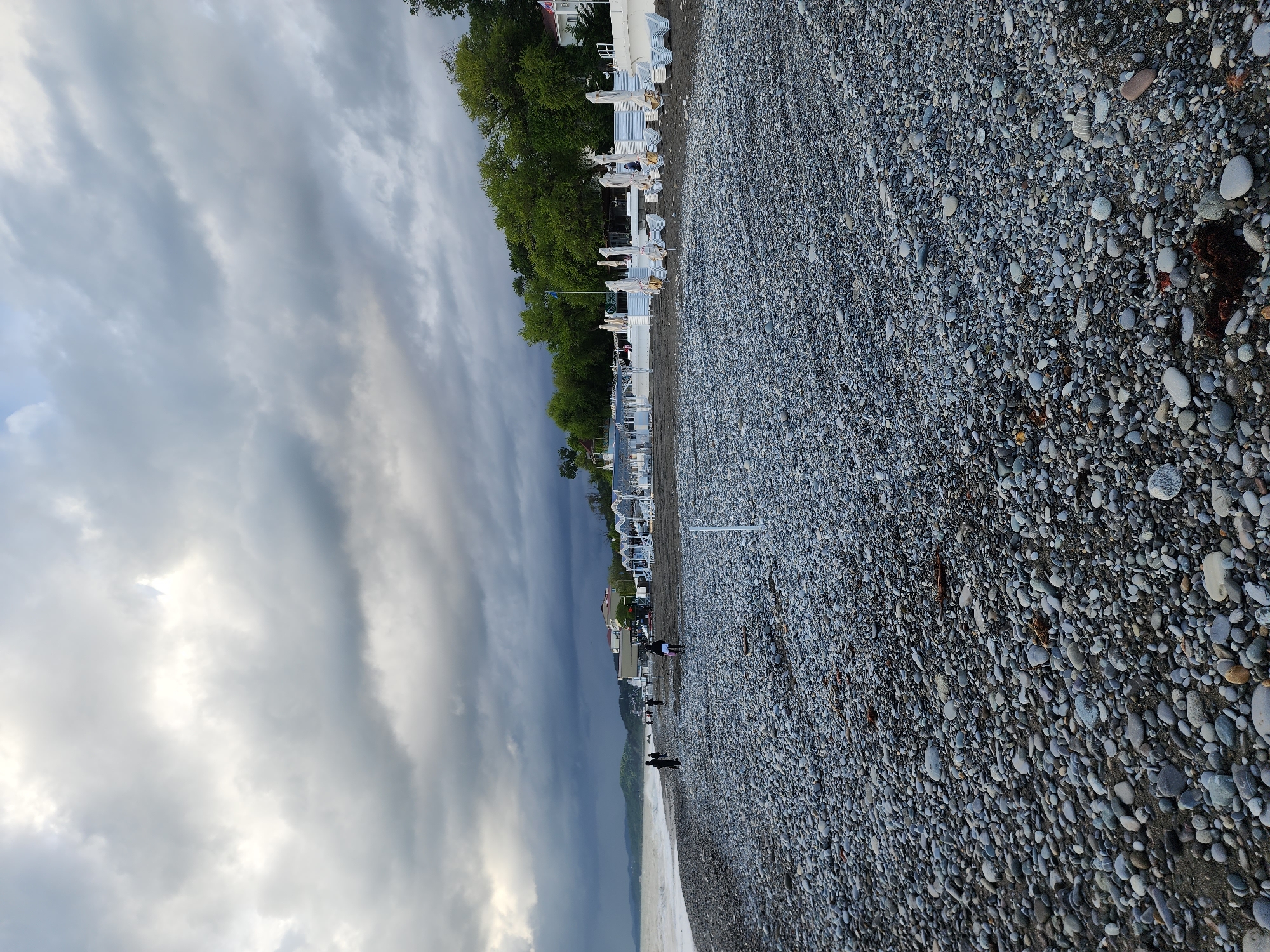 The image shows a pebbly beach on a cloudy day, likely in Sochi, Russia. A line of white beach umbrellas and lounge chairs extends along the upper edge of the beach, bordered by trees. Several people are visible walking near the shoreline. The sky is filled with dark, overcast clouds. The beach is covered with small pebbles of various sizes and colors. The sea is visible along the bottom edge of the frame, with a small wave breaking.