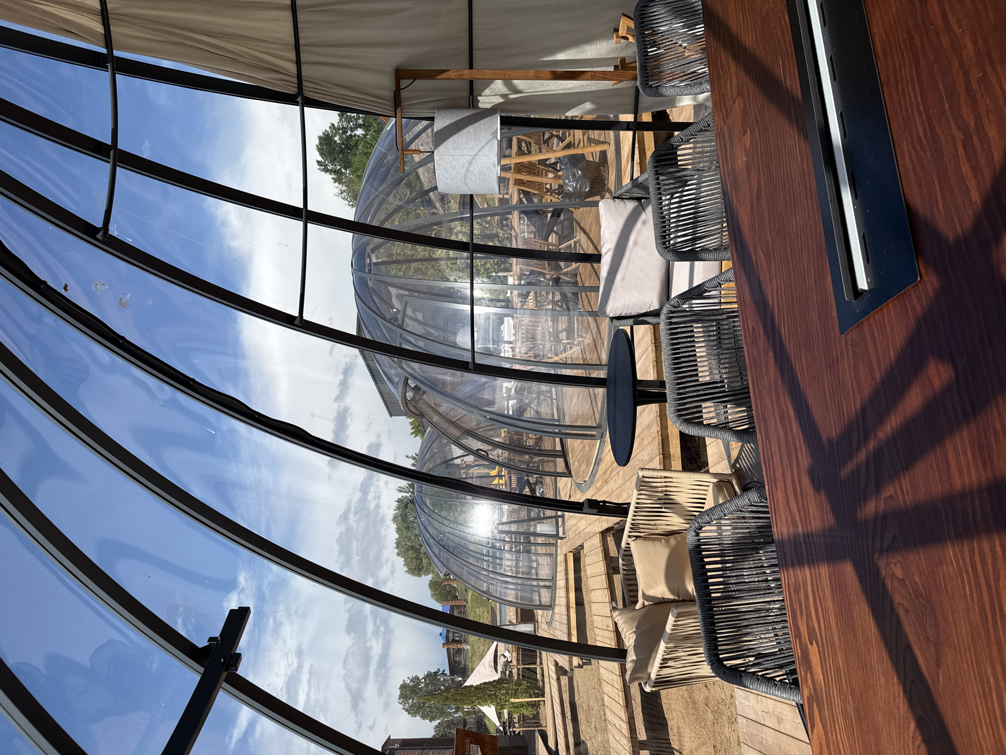 The image shows a semi-outdoor dining area in Votkinsk, Russia. The space is enclosed by a geodesic dome made of transparent panels and dark metal frames, offering views of the sky and outdoor scenery. Inside the dome, there is a wooden deck serving as the floor. The dining area features a long, dark wooden table with built-in lighting, surrounded by woven chairs with light-colored cushions. Other similar domes are visible in the background. The overall setting appears to be a restaurant or event space with a contemporary design.