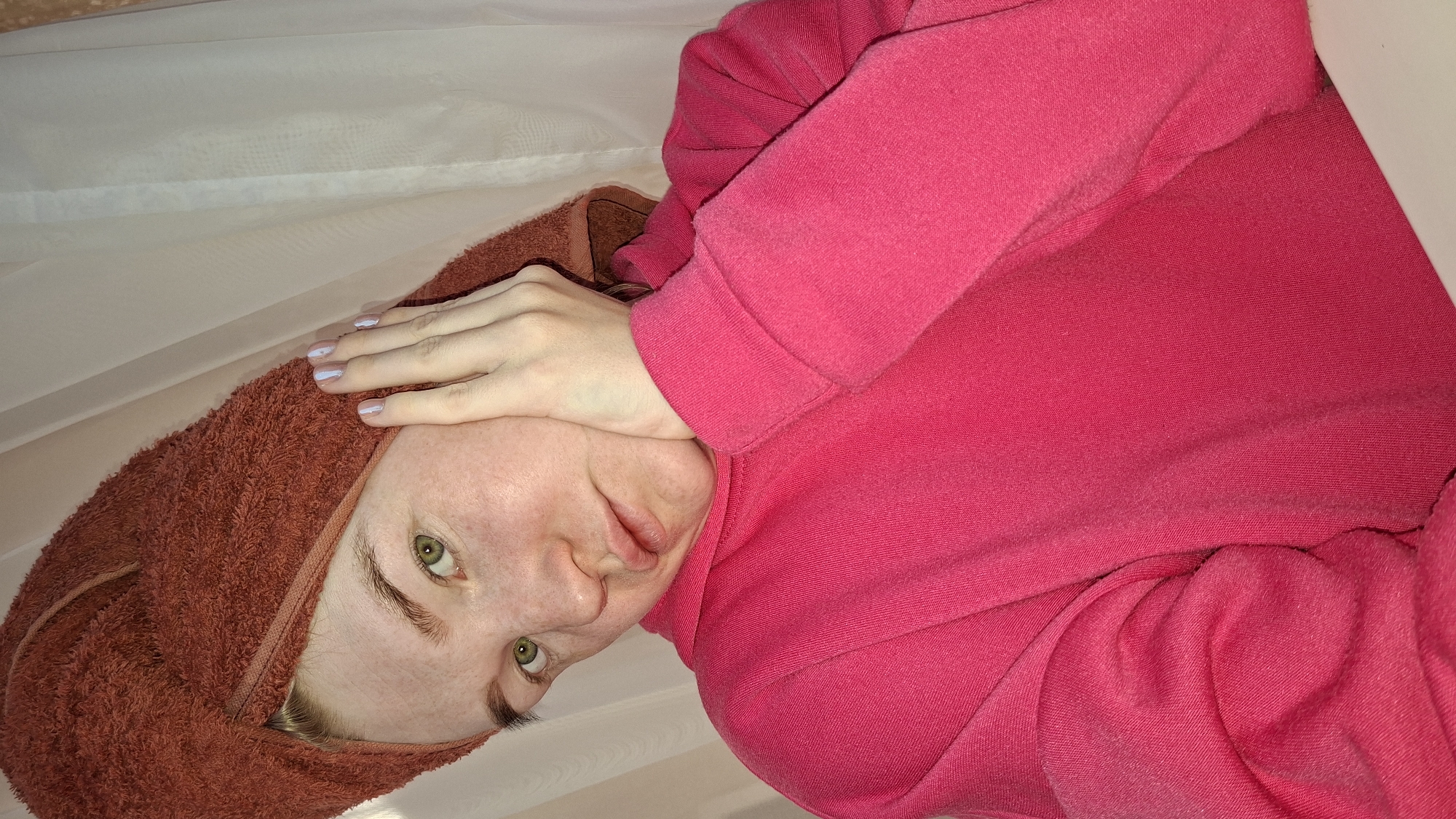 The image displays a headshot of a person, likely a young adult female, captured from an overhead perspective and rotated onto its side. The subject has fair skin, prominent green eyes, and light brown eyebrows. Her expression is neutral, possibly slightly pensive or tired, as she gazes directly at the viewer.
She is wearing a dark pink or magenta long-sleeved hooded top or sweatshirt, which occupies a significant portion of the lower right frame. A rust-orange towel is wrapped around her head, resembling a turban or hair wrap, suggesting she might have recently washed her hair or is engaged in a post-shower routine. Her left hand is raised to her forehead, touching the edge of the towel. The nails on this hand are painted with a light, possibly white or very pale blue, polish, with a small darker detail visible on some fingernails.
The background is simple and light-colored, appearing to be an indoor setting such as a plain wall or curtain. There are no distinct features in the background to provide specific location cues other than the general context of being indoors. No text is visible in the image. The lighting is soft and even, without harsh shadows, indicating an ambient indoor light source. The overall impression is a casual, intimate portrait.