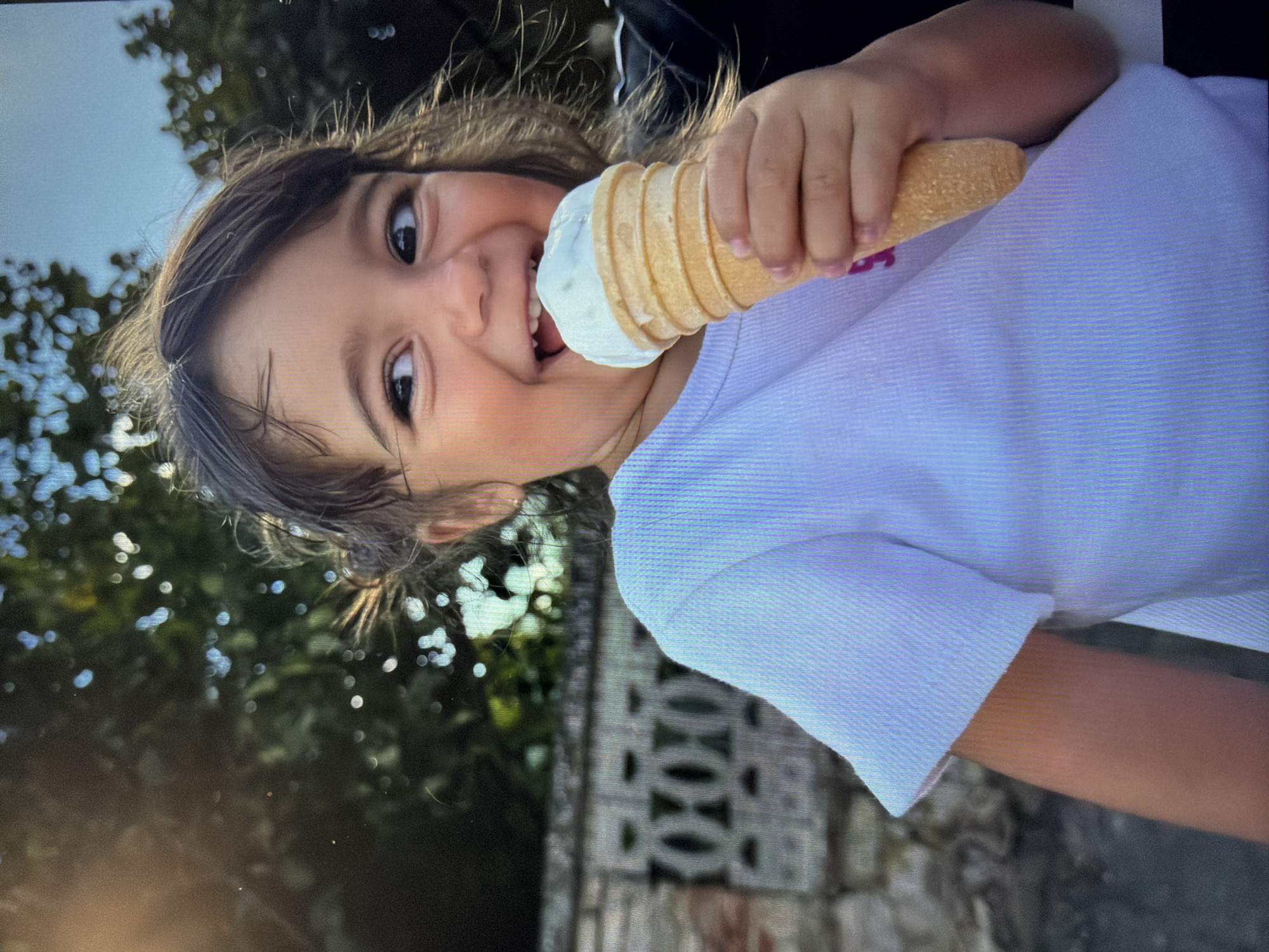 A young girl is eating an ice cream cone outdoors. The cone is tan with vanilla ice cream. She is wearing a short-sleeved white shirt. In the background is greenery and a brick wall with a geometric pattern. The location is Burgas, Bulgaria.
