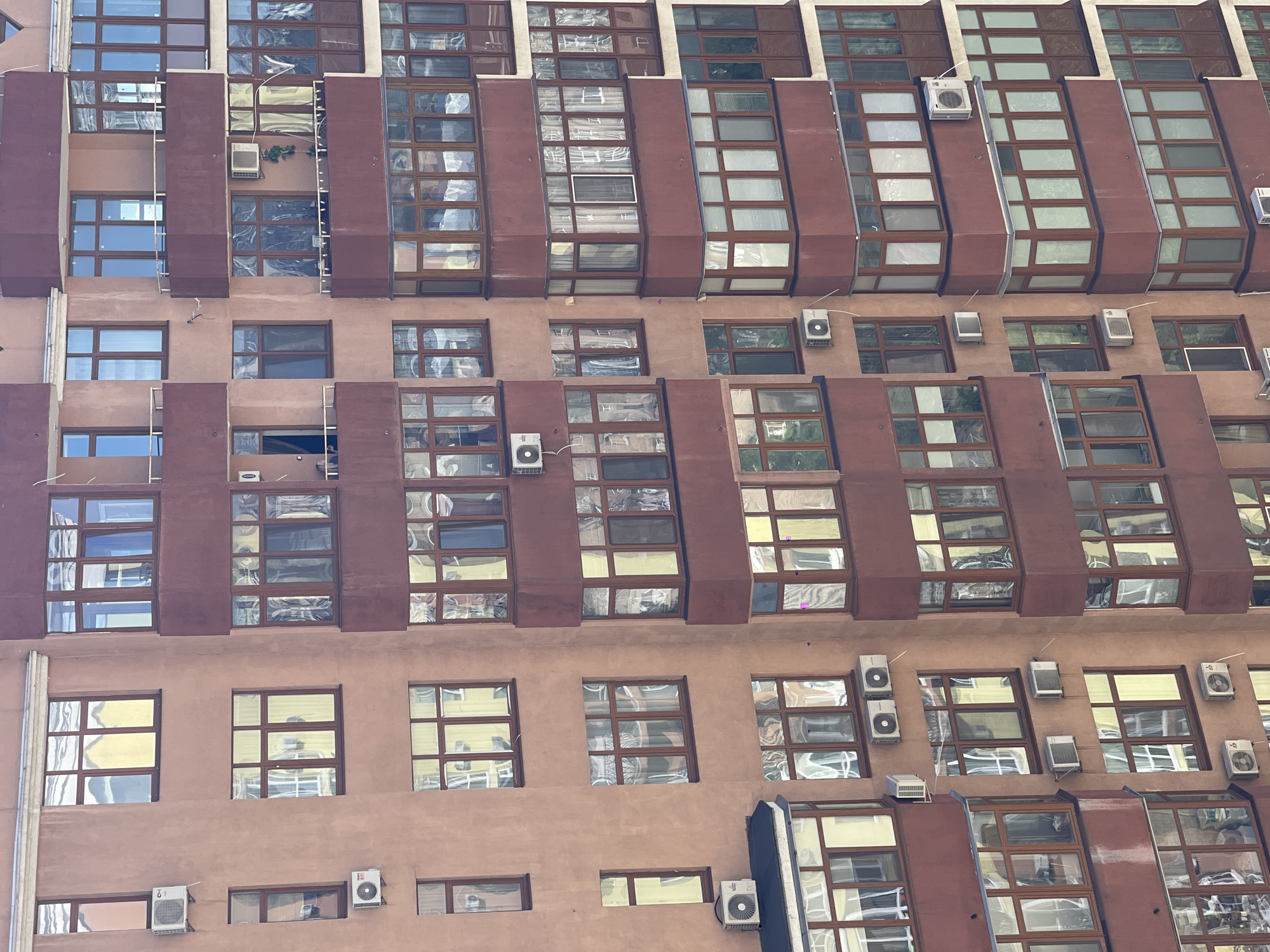 The image is a close-up, straight-on shot of a section of a modern building in Kyiv, Ukraine. The building's facade is predominantly a muted pinkish-brown color, punctuated by numerous windows framed in dark brown. The windows are of varying sizes and configurations, some featuring a grid-like pattern of smaller panes, while others are large, rectangular openings. Many of the windows have a reflective quality, showing distorted reflections of what appears to be the sky or other buildings.
The architecture features protruding elements and angled surfaces, creating a dynamic and somewhat angular appearance. On some of these protruding sections, and on the flat parts of the facade, several white air conditioning units are visible, mounted externally. The design suggests a commercial or residential building with a contemporary aesthetic.
There are no people clearly visible in the image, nor is there any obvious activity or event taking place. The lighting suggests it is daytime, with no strong shadows indicating a specific time of day. The weather appears to be fair, as the sky is not overcast. The overall impression is one of urban architecture, with a focus on the repetition of windows and the functional additions of air conditioning units. No visible text is present in the image.