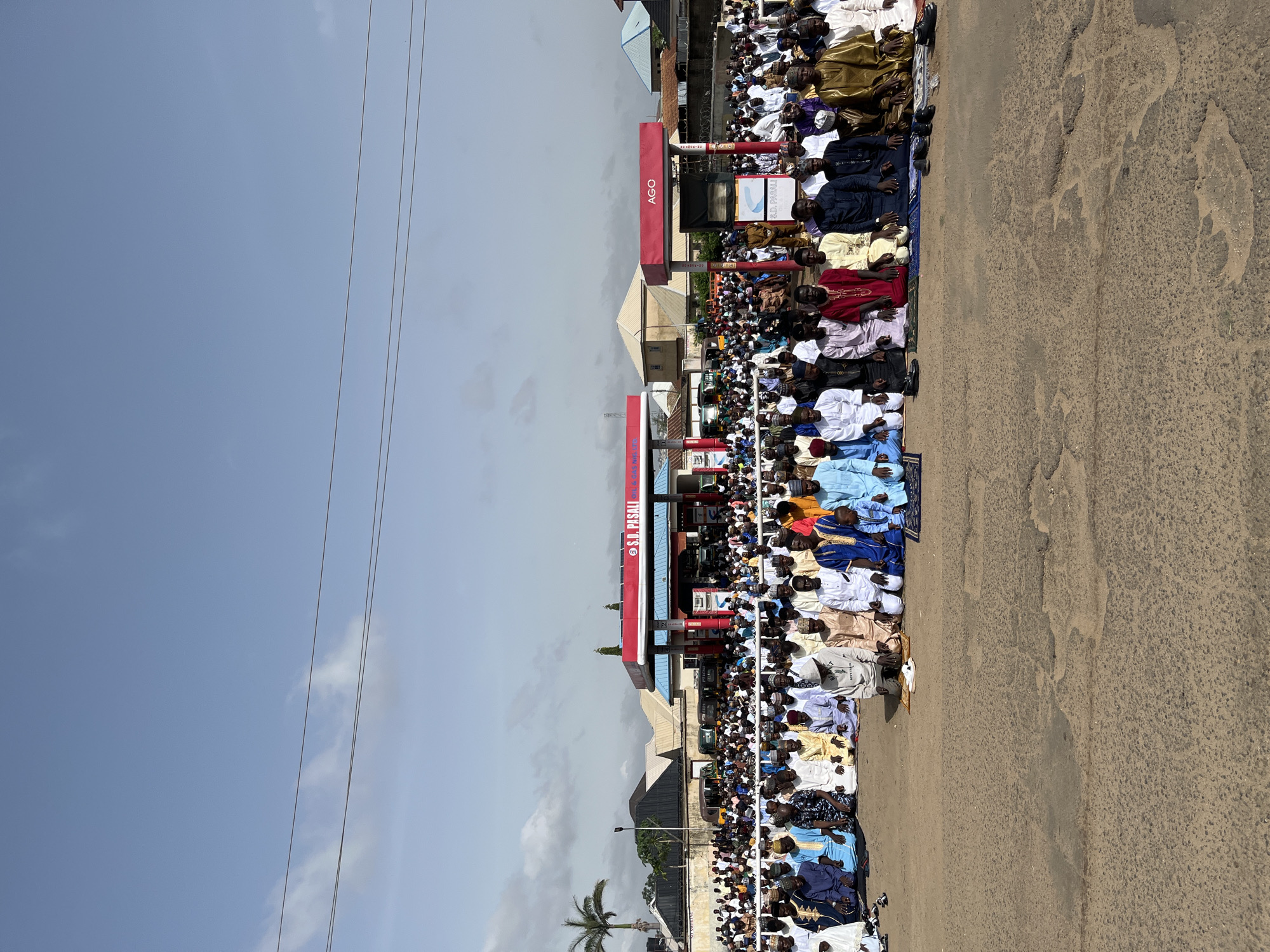 A large group of people are kneeling on the side of a street. They are lined up in rows, facing away from the viewer, and are arranged on mats or rugs. Many are wearing traditional African attire in a variety of colors, including blues, whites, yellows, and golds. A gas station is visible in the background with the brand name "AGO" prominently displayed. In front of the gas station is a sign that says "S.A. PASALI." The road is rough and worn, and the sky is a light blue with some visible power lines. A palm tree is visible near the bottom of the frame. The city is unknown.