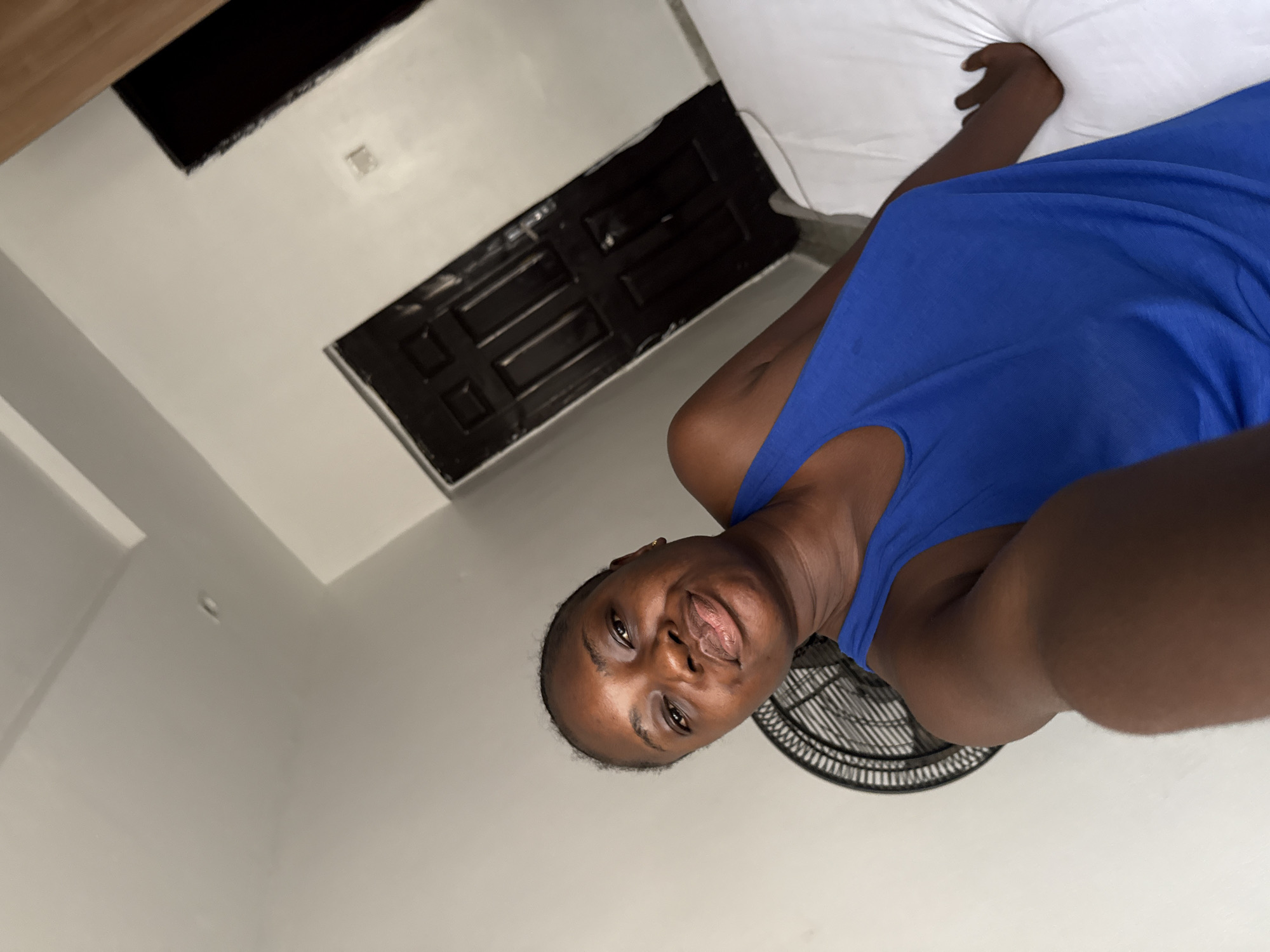 A selfie of a dark-skinned woman in a blue sleeveless top. She is indoors, possibly in a room with white walls. A black window is visible in the background. A white pillow is present in the upper right corner of the frame. A black fan is located in the right lower corner of the frame. The location is Iddo Tudunwada, Nigeria.
