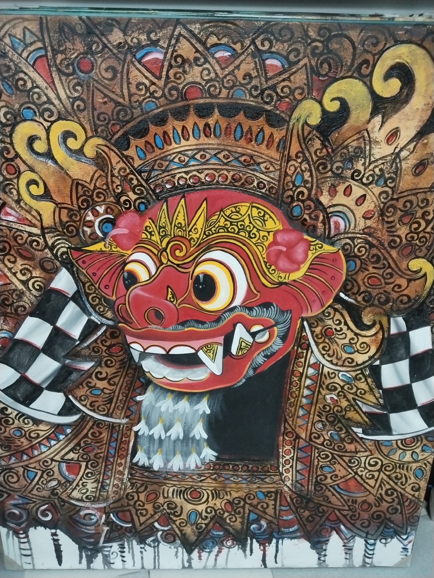 The image is a close-up, eye-level photograph of a vibrantly painted Balinese Barong mask. The mask, predominantly red, features large, round, white-and-yellow eyes with black pupils, sharp white teeth, and a prominent snout. It is adorned with intricate yellow and gold ornamentation on its forehead and sides. A bright red flower is visible on the right side of its head. The mask is framed by a backdrop of elaborate, stylized patterns in gold, brown, black, and red, with accents of blue and white. Two checkered black and white flags are positioned on either side of the mask. Below the mask, a suggestion of a grey, furry or feathered neck is depicted, and the lower portion of the artwork shows more detailed decorative patterns with dripping paint effects at the bottom. The style is indicative of traditional Balinese art, often found in cultural centers like Kuta, Bali, Indonesia. The context suggests this is a piece of art, likely a painting or a relief, displayed in a setting that showcases Balinese cultural heritage, potentially in a gallery, shop, or public space within Kuta.