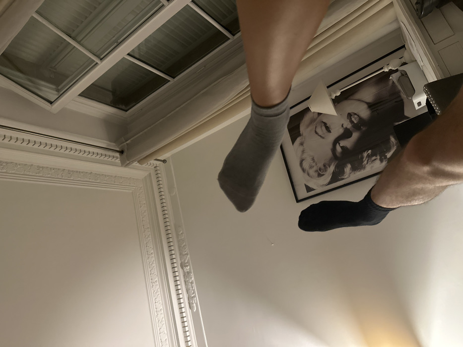 A low-angle shot of a room's interior shows a person's legs with mismatched socks; one gray and one black. The legs are positioned in front of a framed picture of Marilyn Monroe and a white wall. Above, a skylight with a decorative white frame and blinds is visible. A curtain rod and ornate wall molding also appear in the frame, suggesting a traditionally styled room. The shot is taken from a perspective looking upwards.