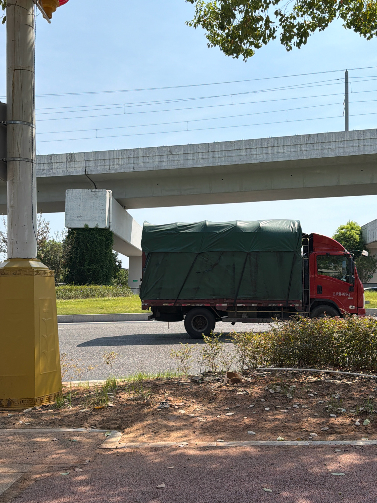 A red truck with a green tarp covering its cargo is driving on a road under an elevated concrete structure. The truck has visible text on its side, including "总质量4495KG" and "165". To the left of the truck, a tall, yellow, cylindrical structure with decorative markings stands near the roadside. Trees and greenery are visible in the background, along with a clear blue sky. The scene appears to be in a part of Jiangjin, China, potentially near a transportation artery or infrastructure.