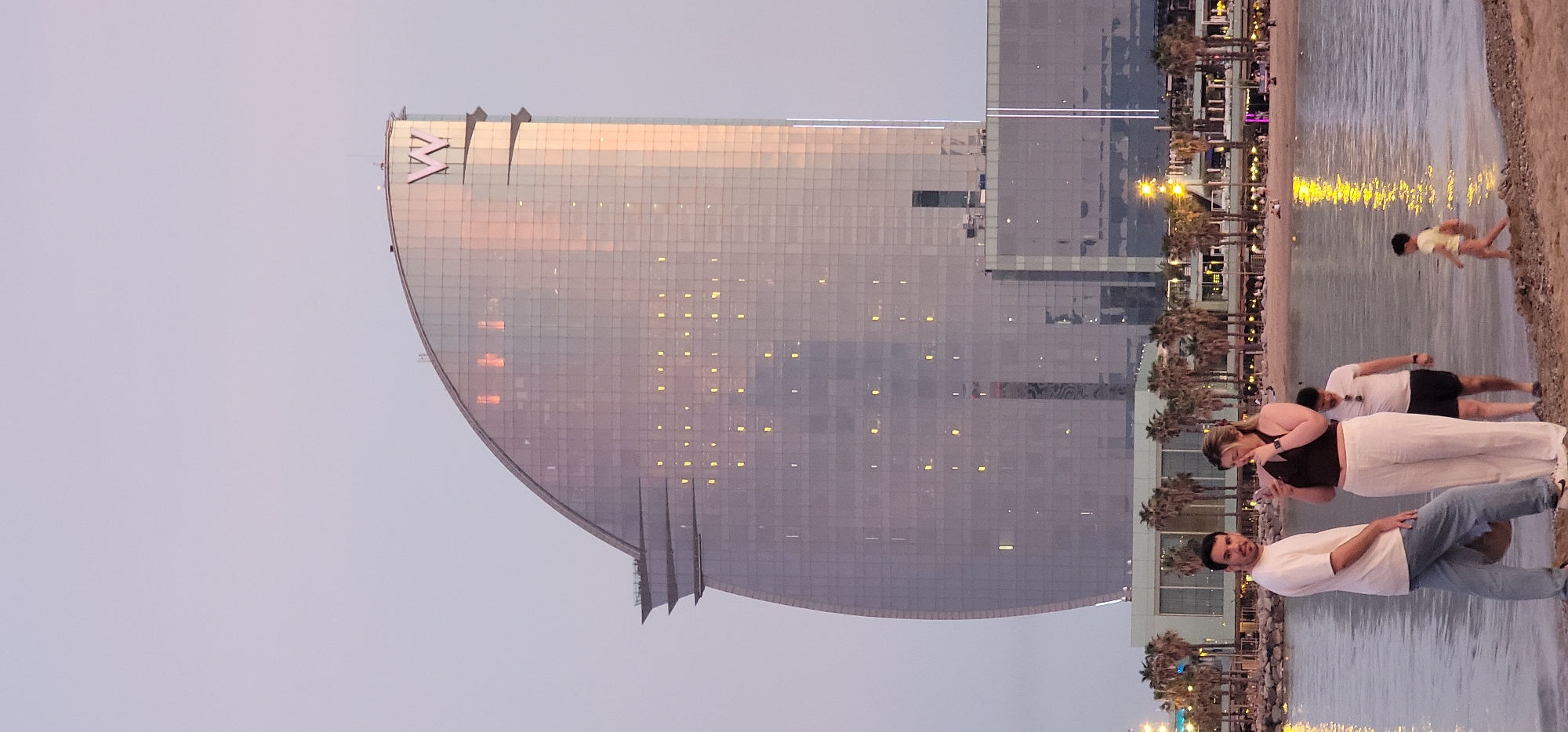 A horizontal shot depicts a beach scene in Barcelona, Spain at dusk. The W Barcelona hotel, a modern architectural landmark with a distinctive sail-like design, is positioned to the left of the frame. Its glass facade reflects the sunset colors. In the foreground, several individuals are present: a man in a white shirt and jeans standing near the water's edge; a woman wearing a dark top and white pants, holding a cell phone; and a man with dark shorts and a white shirt lying on the sand. A child in a yellow shirt is seen running towards the water. Palm trees line the beach behind the people, and streetlights illuminate the area. The sea reflects the lights from the buildings, enhancing the scene's ambiance.
