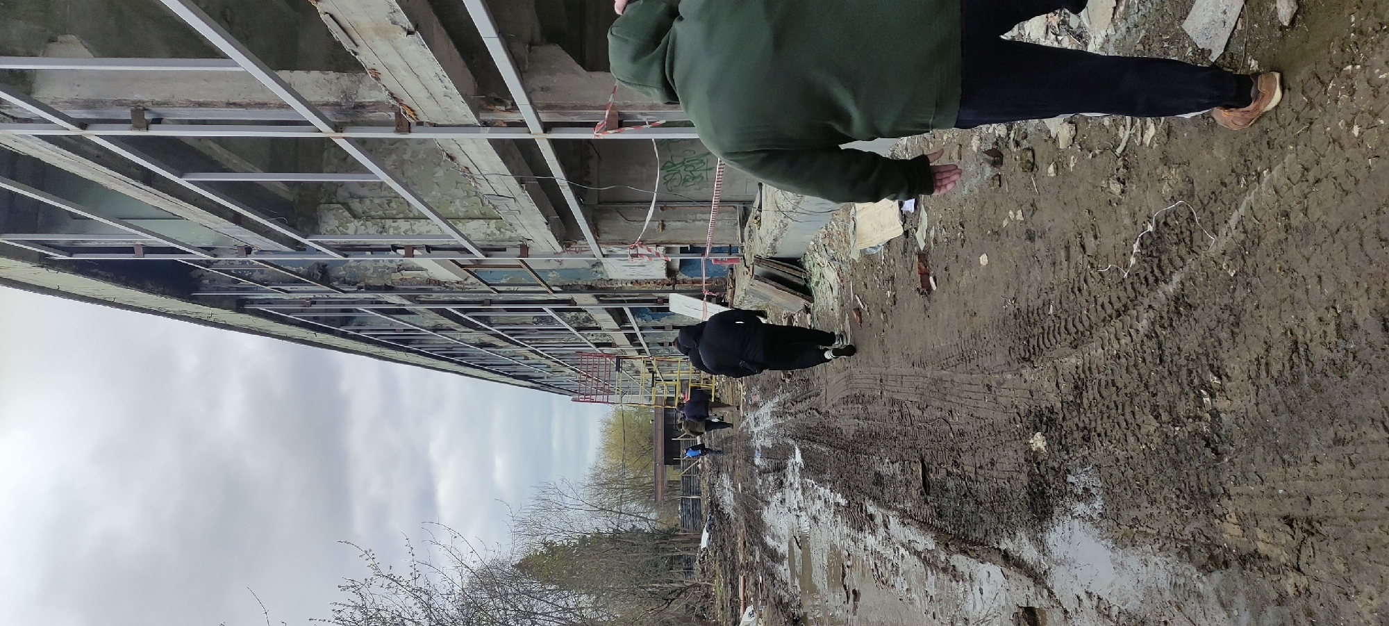 The image depicts an abandoned building site in Fryazino, Russia, under an overcast sky. The structure, with its exposed framework, suggests a state of disrepair or ongoing demolition. Several individuals are present. One person, wearing a green jacket and dark trousers, stands near the building, seemingly observing the site. Further along the muddy ground, another person in a black jacket and trousers walks away from the viewer, while additional figures are visible in the distance near the building's perimeter. The foreground is dominated by a muddy path marked with tire tracks, indicating vehicle movement and heavy activity. Bare trees and vegetation are visible along the edge of the site, adding to the bleak, post-industrial atmosphere.