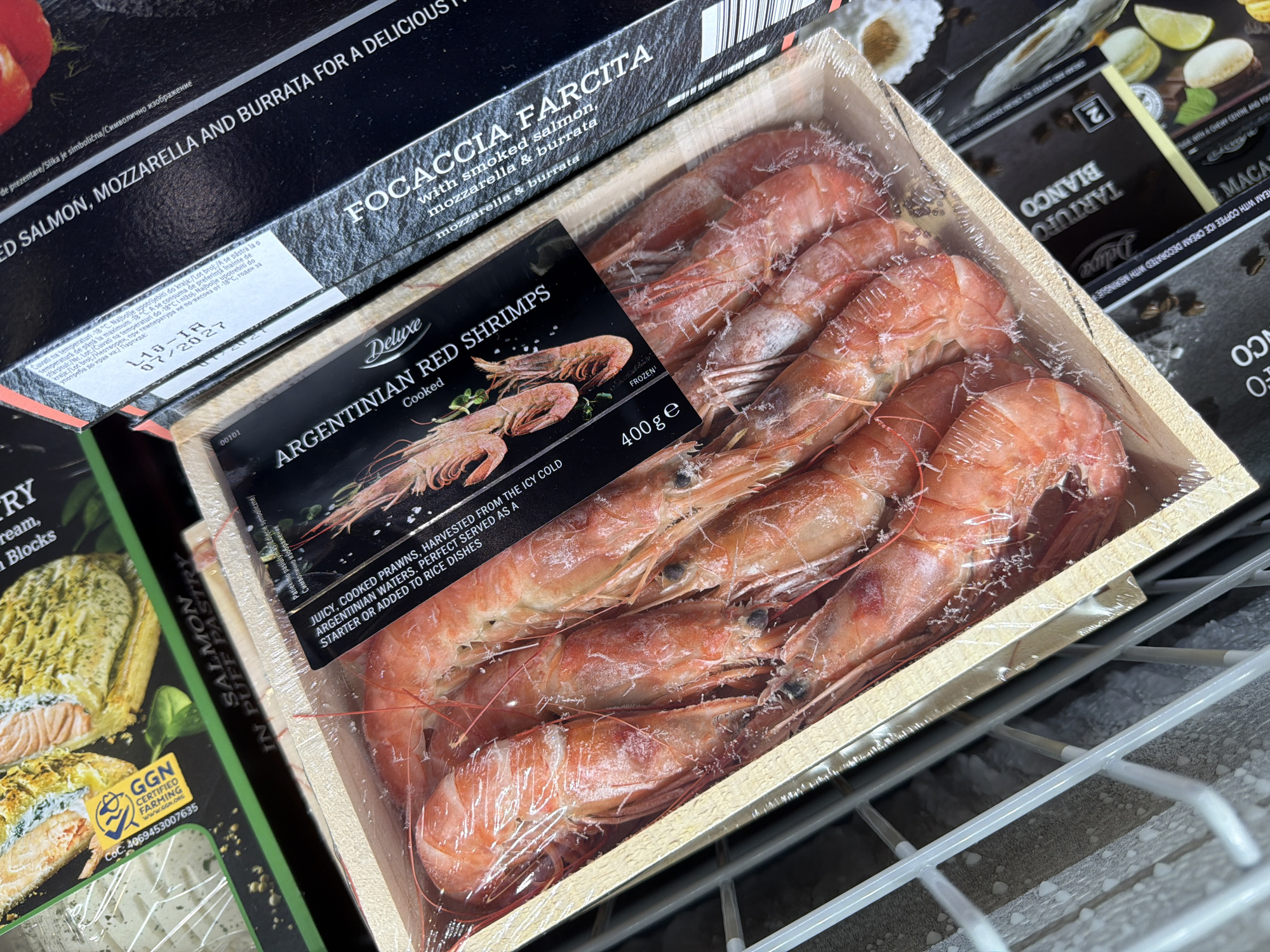 This media file captures a close-up view of various frozen food products arranged within a supermarket freezer, likely in Pazardzhik, Bulgaria, given the provided context. The primary focus is on a transparent plastic-wrapped tray of "Deluxe Argentinian Red Shrimps," which are cooked and presented as juicy prawns harvested from icy cold waters, suitable as a starter or added to rice dishes. The package clearly states "FROZEN" and contains "400g e" of product. The shrimps themselves are reddish-orange and coated with a thin layer of frost, indicating their frozen state.
Surrounding the shrimp package are other product boxes. Partially visible to the upper left is a black box labeled "FOCACCIA FARCITA with smoked salmon, mozzarella & burrata," which also shows storage instructions in multiple languages, including Bulgarian, indicating storage at -18°C and a best-before date of "07/2027." Another product visible to the lower left is a green and black box featuring "SALMON IN PUFF PASTRY" and a "GGN CERTIFIED FARMING" logo with a CoC number. To the upper right, parts of a "DELUXE TARTUFO BLANCO" (white truffle) product and "MACARONS" are also visible.
The setting is characterized by the frosty metal grates of a freezer shelf, suggesting a cold supermarket environment. The packaging designs, particularly the "Deluxe" branding, imply premium food items. No people are visible, and the scene depicts a static display of goods ready for purchase.