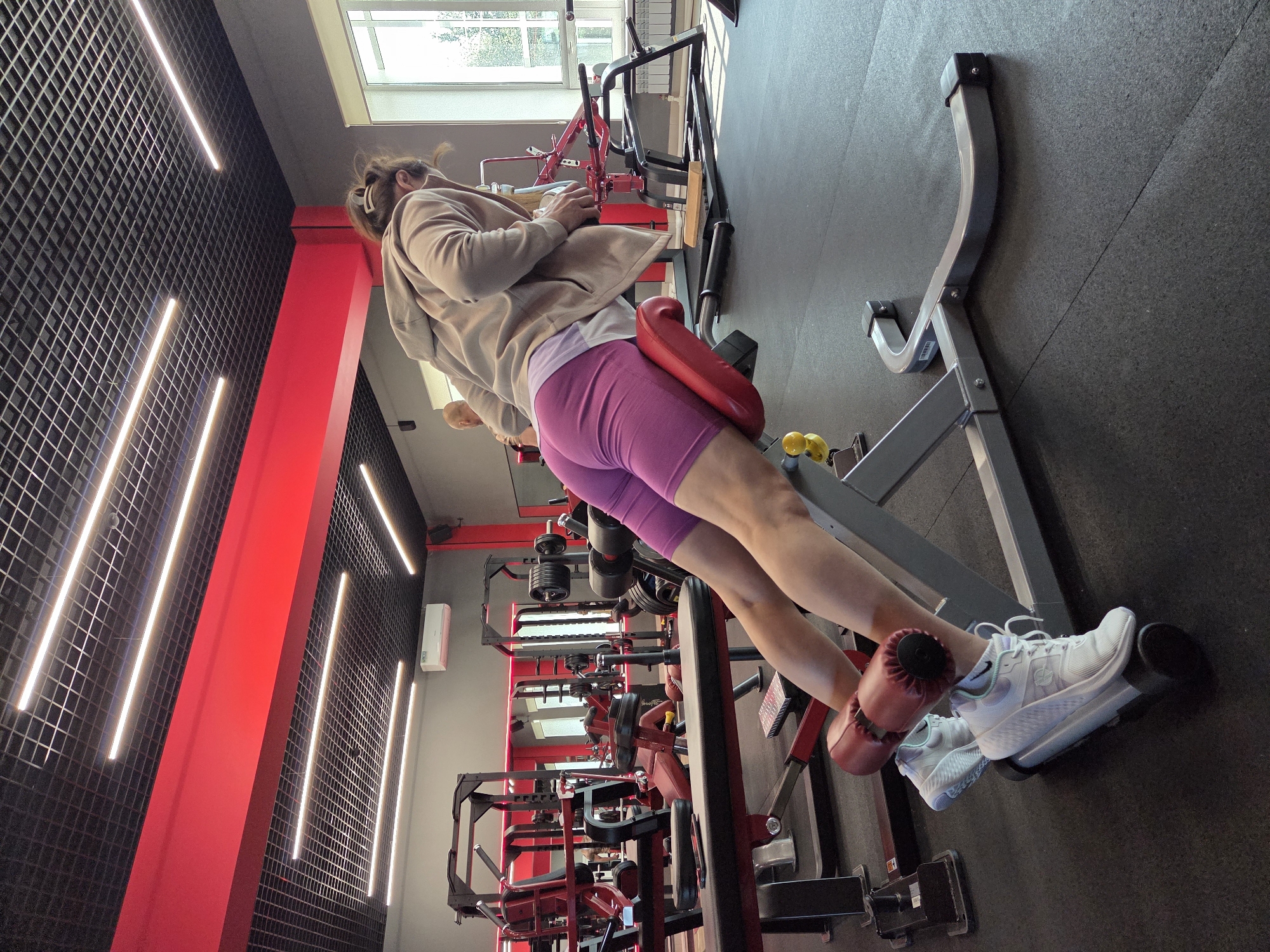 The image depicts the interior of a gym, likely located in Birobidzhan, Russia, given the context. A woman is engaged in a glute-ham raise exercise on a red and grey piece of equipment. She is positioned with her legs secured by padded rollers and her torso extended forward, supported by a padded bench. She is wearing a light beige hooded sweatshirt and bright purple athletic shorts. Her white athletic shoes are visible, secured to the machine's footplate. The gym environment includes other exercise machines, weight racks with dumbbells, and a mirrored wall in the background, all contributing to a typical fitness facility setting. The ceiling features a black grid with integrated LED lighting strips and a prominent red beam. A window with natural light is visible to the right of the frame, and an air conditioning unit is mounted on the wall. The flooring is black rubber gym matting.