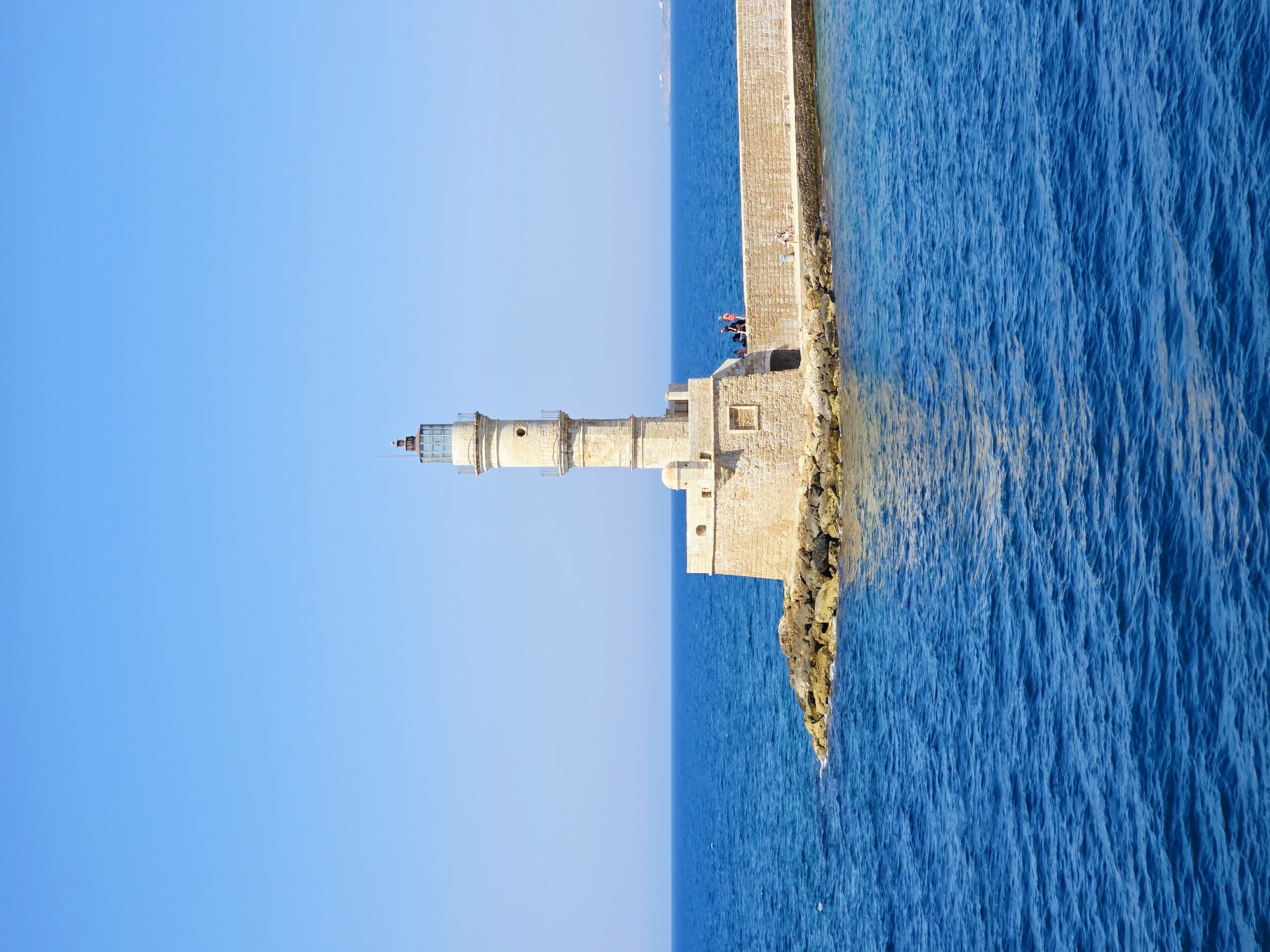 This image captures the Venetian Lighthouse of Chania, a prominent landmark in Chania, Greece, situated at the end of a stone breakwater. The lighthouse, constructed of weathered stone, stands tall and slender against a clear, bright blue sky. Its octagonal base and cylindrical tower are topped with a lantern room. Several people are visible on the breakwater, some sitting and others standing, appearing to admire the view. The breakwater itself is made of large, beige stones and extends into the deep blue, rippling waters of the sea. The surrounding environment is characterized by the expanse of the ocean meeting the horizon under a cloudless sky, suggesting a sunny and calm day. The image is taken from a slightly elevated perspective, possibly from a boat, looking towards the lighthouse and breakwater.