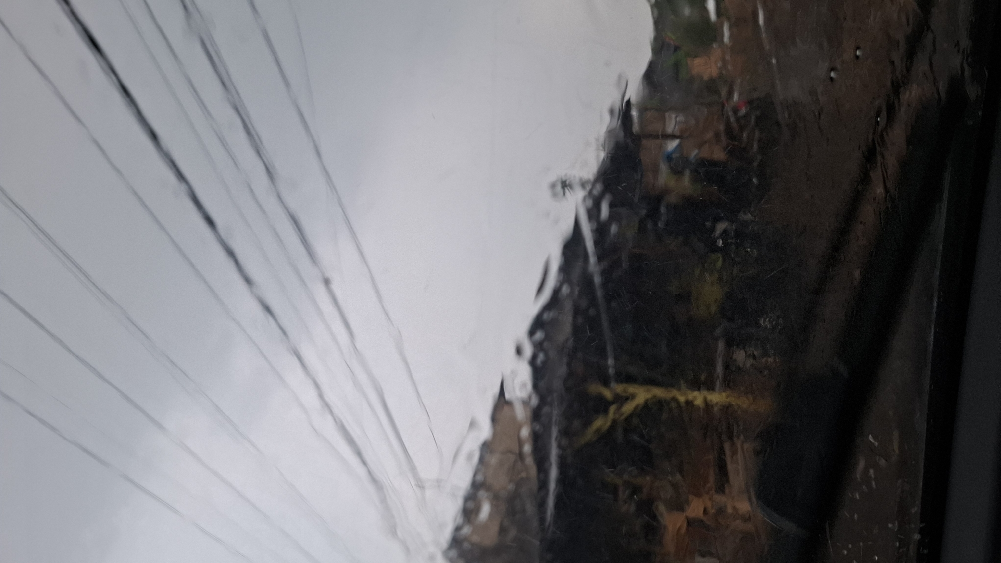 The image, taken from inside a vehicle looking out, depicts a blurry scene of a dwelling or structure with a dark roof, situated on a slope. The environment appears to be a rural or semi-rural area in Cilaku, Indonesia, possibly a roadside view. Several dark power lines crisscross the overcast sky, running diagonally across the upper portion of the frame. The foreground shows a portion of the vehicle's interior, likely the dashboard or windshield wiper. The overall impression is one of a somewhat weathered, possibly impoverished, settlement under a gray sky, with the ubiquitous presence of overhead electrical infrastructure. The image quality is degraded by blur and reflections, suggesting it was captured while the vehicle was in motion or through a dirty or rain-streaked window.