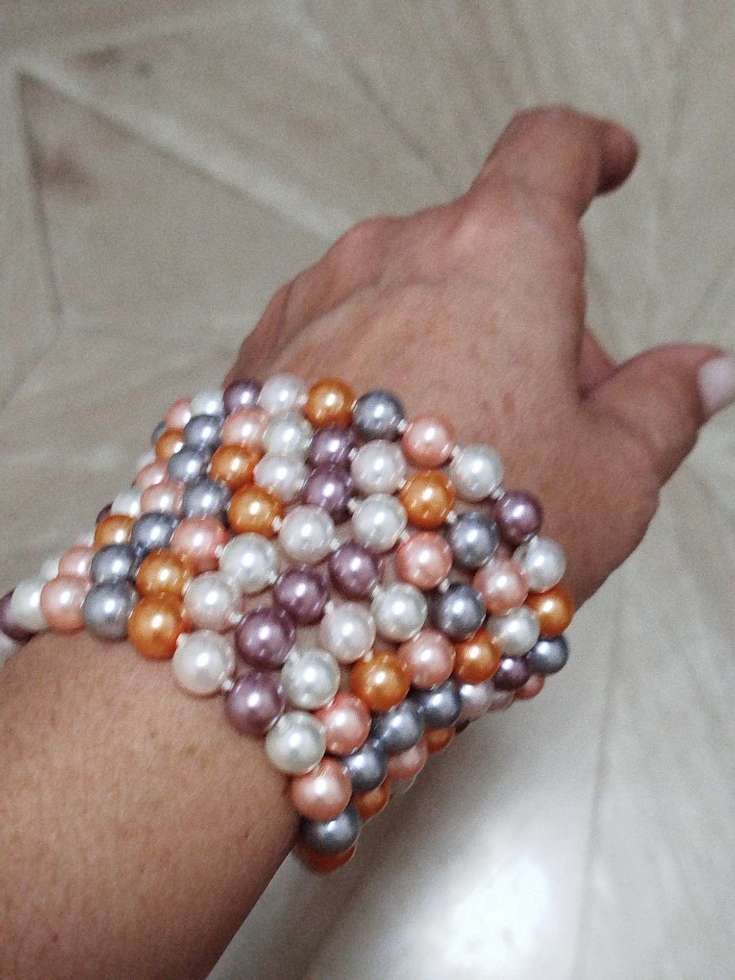 A close-up, slightly angled shot shows a multi-strand beaded bracelet on a person's wrist. The bracelet is made of pearls in shades of white, light pink, orange, and gray, arranged in a layered, flexible structure. The beads are round and have a lustrous finish. The background is blurred and appears to be a neutral-colored, textured surface, possibly a floor or fabric. The image is focused on the bracelet and the lower portion of the hand and wrist, with the rest of the person's arm and body out of frame. The lighting is soft and diffused. There is no discernible connection to Qarağandı city administration, Kazakhstan.