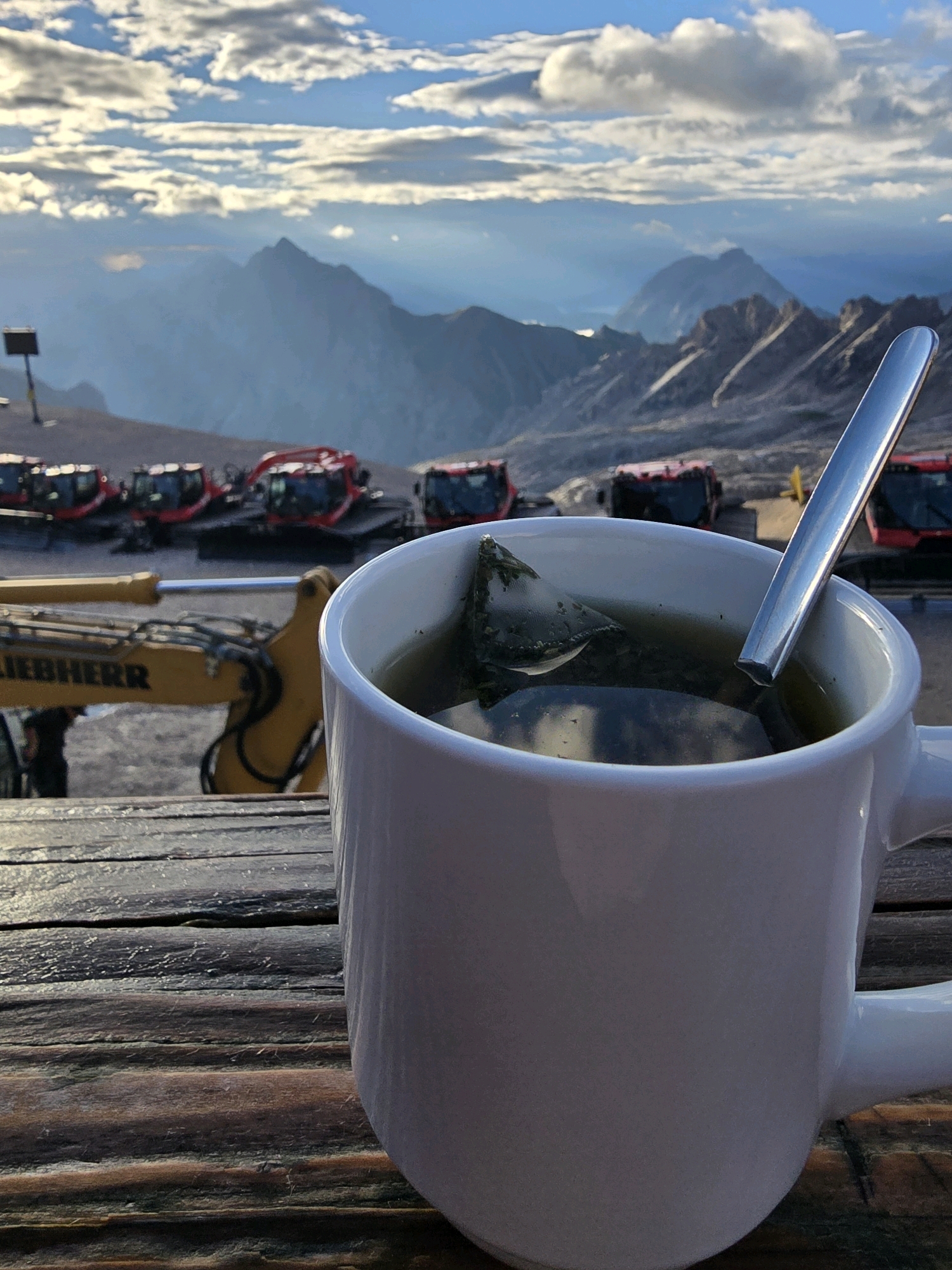 A white mug of tea with a tea bag steeping inside sits on a weathered wooden table. The mug is in the foreground, obscuring part of the background. Behind the mug, a yellow excavator with the brand name "LIEBHERR" visible on its arm is parked. Further in the background, a row of red snow groomers are lined up. Beyond the vehicles, a vast mountain landscape unfolds under a dramatic sky with clouds. The scene suggests a location in a mountainous region, possibly near a construction or maintenance site. Given the presence of snow groomers, it is plausible this is a location in Garmisch-Partenkirchen, Germany, known for its alpine environment and ski resorts. No people are visible in the image.