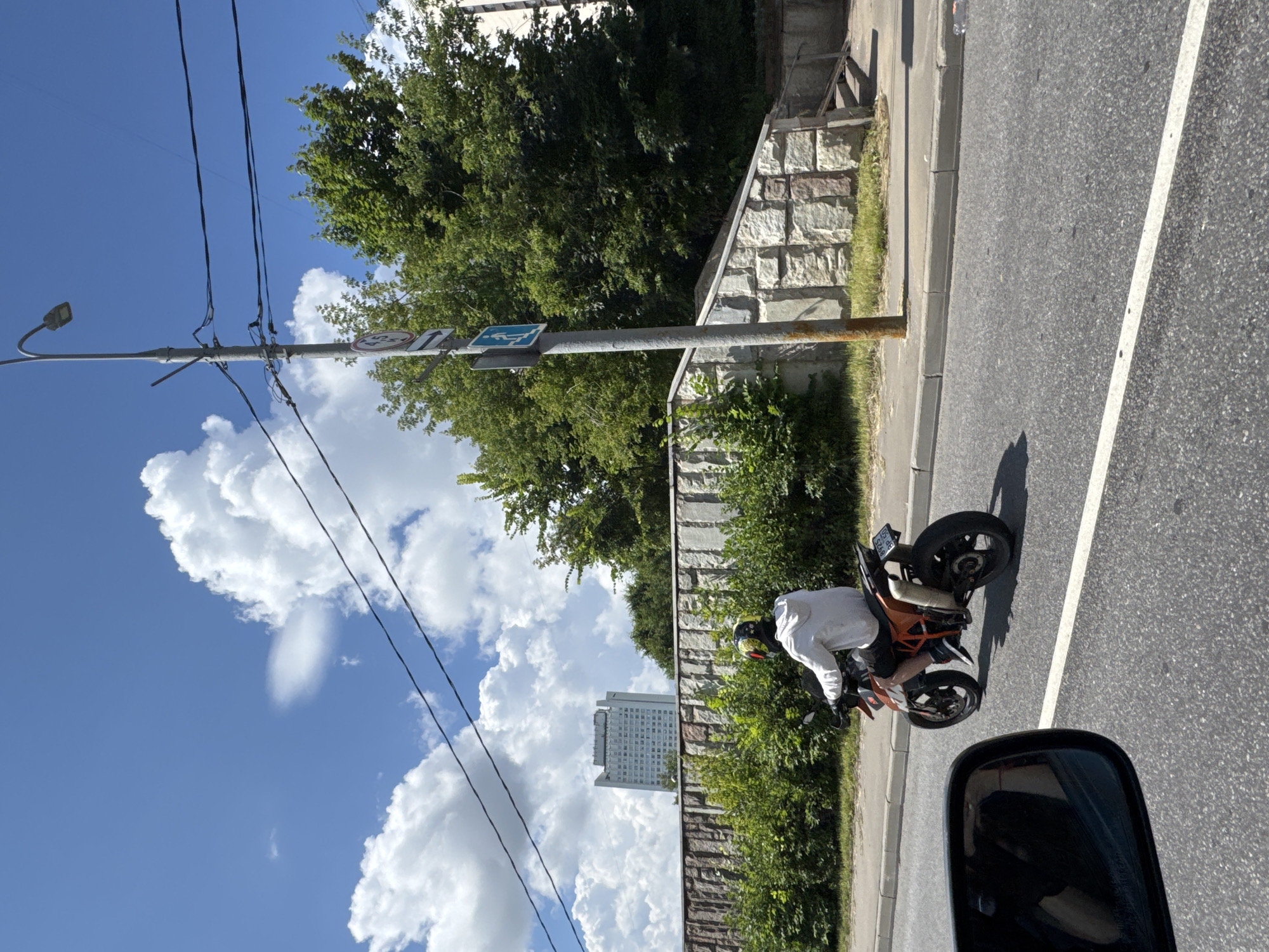 A person wearing a white hoodie and a helmet is riding a KTM motorcycle on a paved road in Zelenograd, Russia. The motorcycle is orange and black with a license plate that reads "4925 BP 150". The rider is seen from the side and slightly from behind, with their legs on the footpegs. In the foreground, a portion of a car's side mirror is visible, reflecting the sky and some greenery. Behind the motorcycle, there is a stone wall with a stepped incline and lush green bushes. Further in the background, a tall, white building with many windows stands against a bright blue sky filled with fluffy white clouds. A lamppost with wires crisscrossing above is also present in the scene.