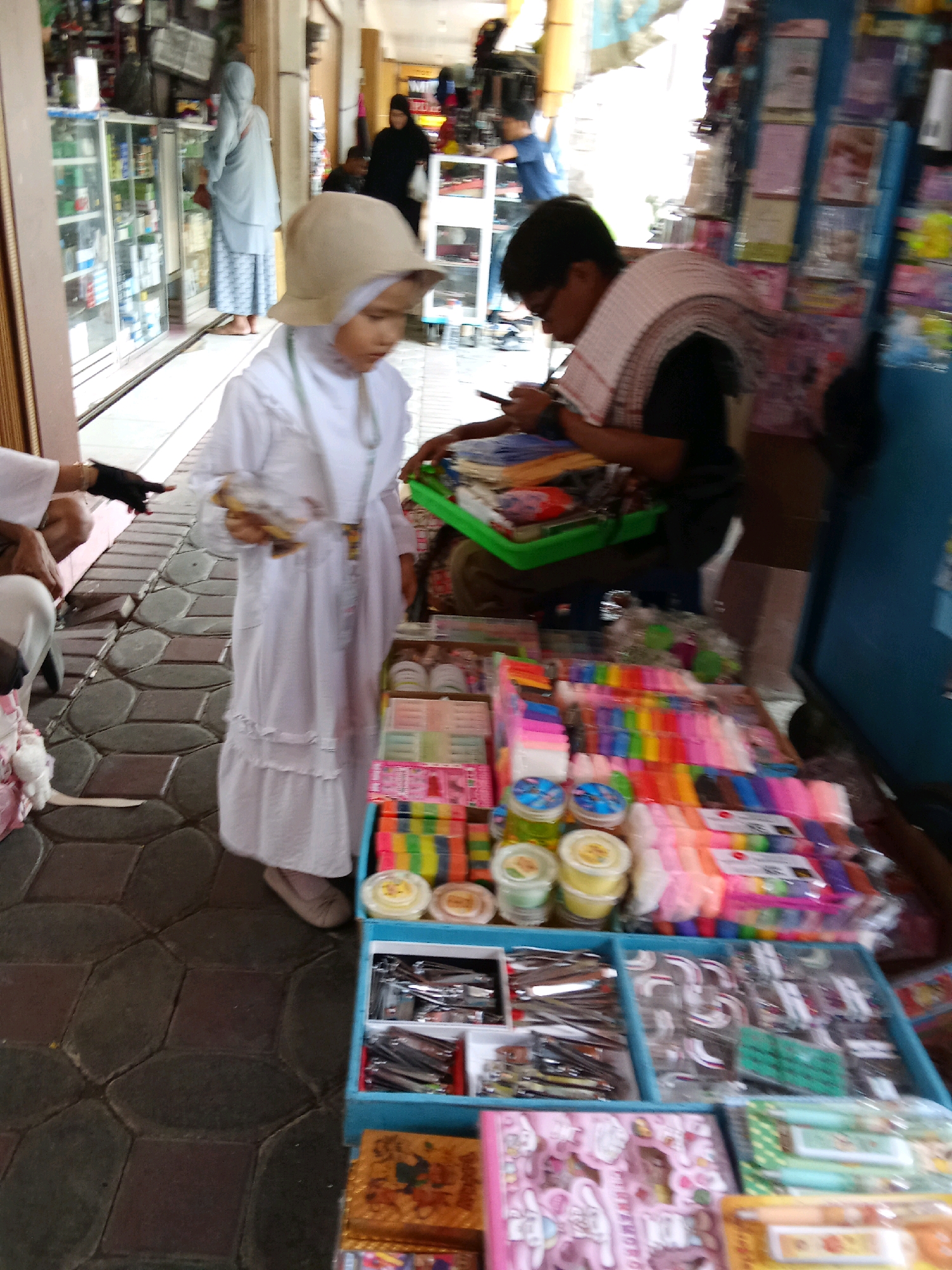 This image depicts a street market scene in Banjarnegara, Indonesia. In the foreground, a young child wearing a white dress and a tan hat is holding a small packaged item and looking down. Beside the child, a stall is displayed with various items for sale, including rows of colorful stationery, jars, and small boxes of items like scissors and other tools. Further back, an adult is seated and looking at a phone, with a patterned scarf draped around their shoulders. In the background, other shoppers and stalls are visible, including a person in a black hijab and another in a light blue outfit. The market appears to be situated outdoors with paved flooring.