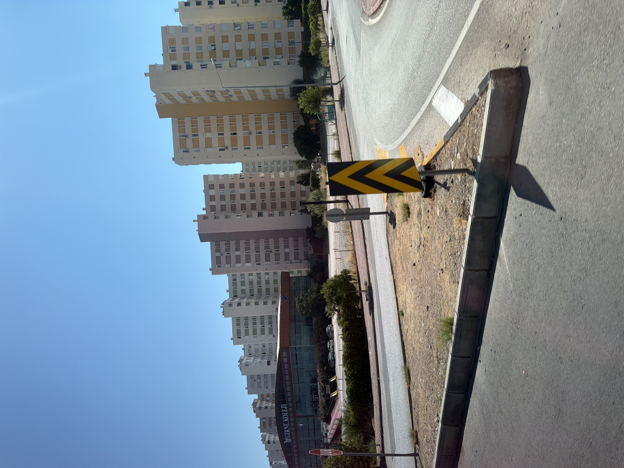 The image captures a street scene in Konyaaltı, Turkey, on a clear, sunny day. In the background, a row of multi-story apartment buildings with light-colored facades and yellow-accented windows stretches across the horizon. In the mid-ground, a modern building with a glass facade prominently displays the text "GİRNE KOLEJİ" (Girne College). The building is surrounded by greenery and a paved area with parked cars. In the foreground, a road curves to the right, bordered by a concrete curb and a patch of dry, sandy ground. A traffic sign with black and yellow chevron markings is positioned on the left side of the road. A red stop sign is visible near the curb on the far left. The sky is a vibrant blue, with no clouds present.