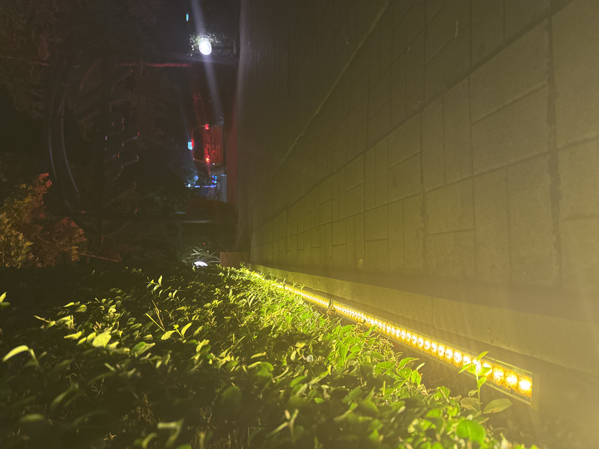 The image depicts an outdoor night scene in Burgas, Bulgaria, characterized by prominent artificial lighting. In the foreground, a vibrant strip of yellow LED lights runs along the base of a textured wall, illuminating the dense, green foliage and bushes in front of it. The wall itself appears to be constructed from light-colored blocks or bricks.
On the left side of the frame, the background is darker, with silhouettes of trees or tall bushes. A large, dark, circular structure, possibly a Ferris wheel or an art installation, is discernible amidst the greenery. Above this, a bright, white light source, resembling a street lamp, emits a strong glow and lens flare. Further in the mid-ground on the left, a vehicle, possibly a public transport bus or a service truck, is visible with distinct red and blue lights.
The time of day is clearly night, with the darkness punctuated by various artificial light sources. The weather appears clear. The primary light source is the warm yellow LED strip, creating a bright and inviting path along the wall, contrasting with the cooler white lights in the distance. The scene evokes a calm, illuminated ambiance in what appears to be an urban or recreational area. There is no visible text in the image.