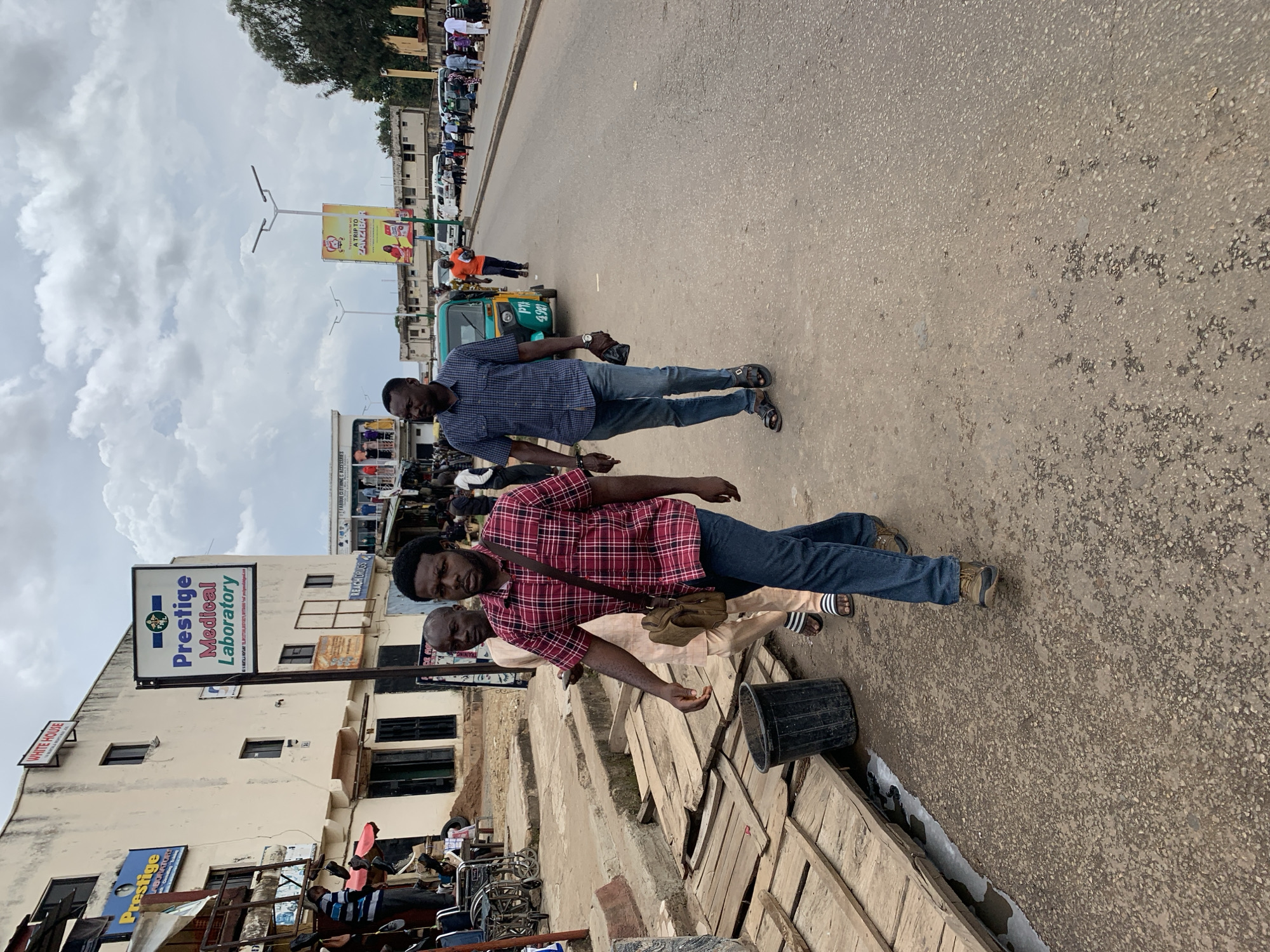 A street scene in Jos, Nigeria, features a cloudy sky and a paved road with a sidewalk. Three men are walking on the sidewalk, and several vehicles, including a green and yellow auto-rickshaw, are visible on the road. Buildings line the street, with one displaying a sign for "Prestige Medical Laboratory." Wooden planks are laid out on the sidewalk, and a black trash can is present. The overall atmosphere suggests a bustling urban environment with commercial activity.