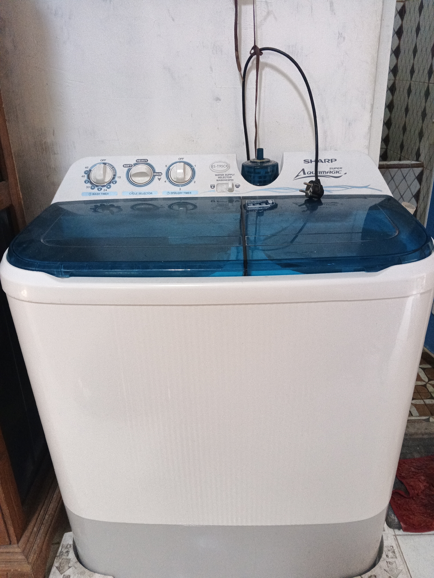 A top-loading, dual-tub washing machine, white with a blue lid, is positioned in a residential setting in Kemangsen, Indonesia. The machine features a control panel with three dials: "Wash Timer," "Cycle Selector," and "Spin-Dry Timer," along with various settings such as "Off," "Heavy," "Soft," and time increments. A black power cord with a plug hangs from above, connected to a blue water inlet valve at the top of the machine. The washing machine is placed on a tiled floor, with its base resting on what appears to be a damaged piece of cardboard. To the left, a portion of a wooden piece of furniture is visible. The background shows a textured, off-white wall and a decorative tiled wall section on the right. A red rug is partially visible on the floor to the right of the washing machine.