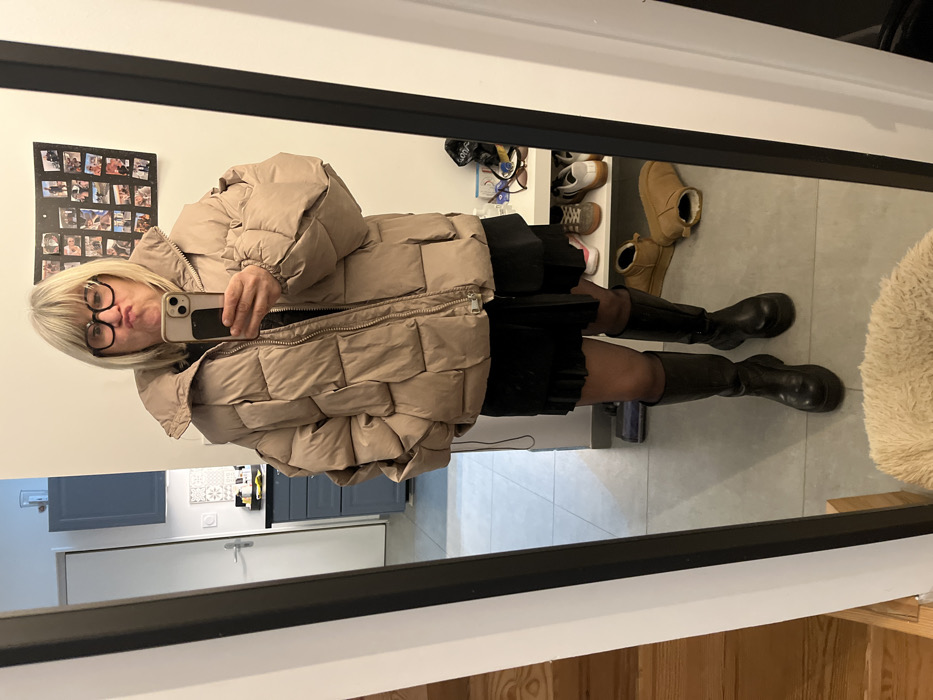 A blonde woman in glasses poses in a mirror, wearing a beige puffer coat, black skirt, and knee-high black boots. Behind her, a shoe rack holds various sneakers and tan boots. A picture collage hangs on the wall above, and reflections show part of a kitchen with gray cabinets and a white door. The floor is a mix of tile and wood.
