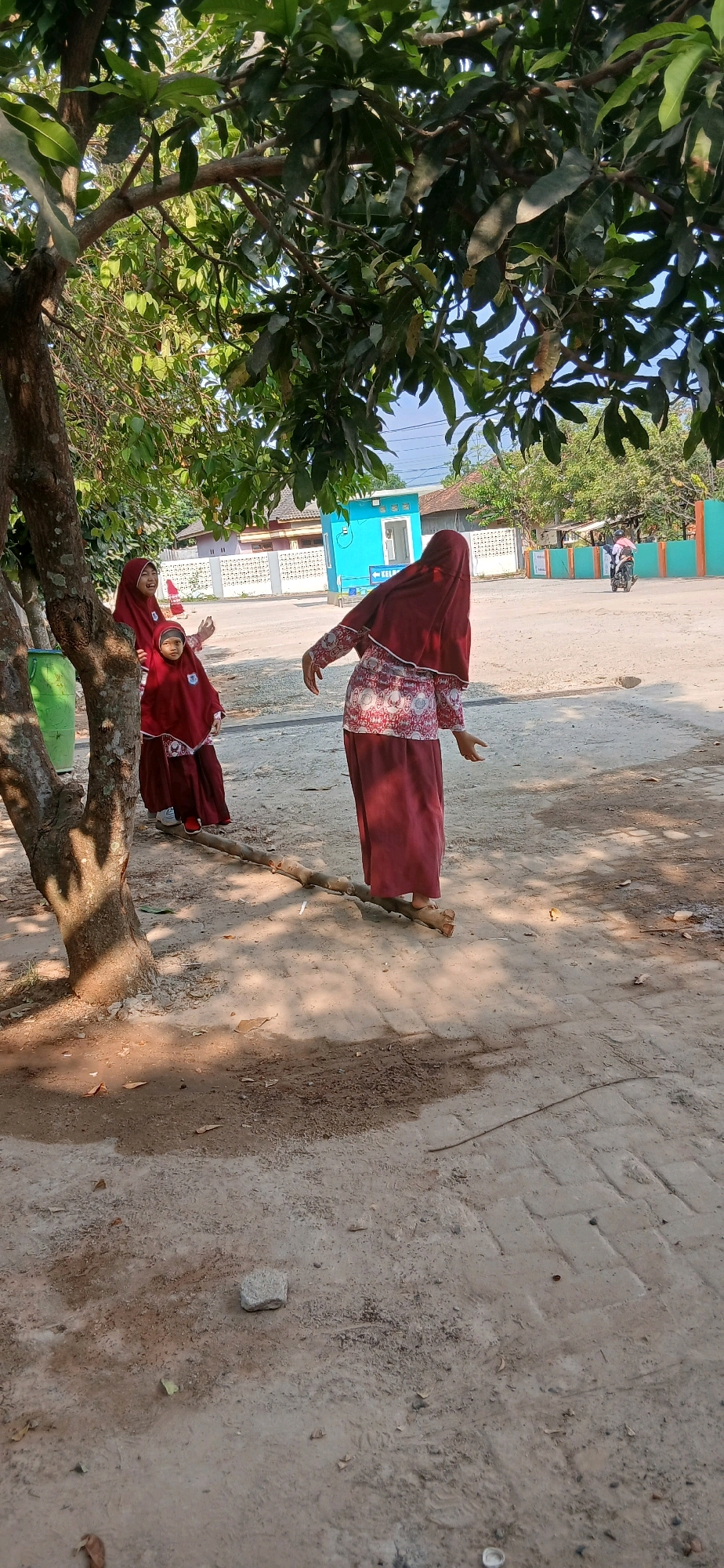 The image depicts an outdoor scene with three individuals, two of whom are girls wearing maroon hijabs and matching long dresses. The third individual, also wearing a maroon hijab and dress, is positioned in the foreground, facing away from the camera. The girls in the background appear to be interacting, with one pointing and the other looking towards the foreground. They are standing under the shade of a tree with green leaves.
In the background, there is a dirt path or road. A blue building with white trim is visible, along with a white fence. To the right, a person on a motorcycle with a pink umbrella is seen riding away. The ground is a mixture of dirt and what appears to be some paved sections. Scattered leaves and small stones are visible on the ground. The scene suggests a casual, everyday setting. This image was captured in the city of Metro, Indonesia, likely during daylight hours, indicated by the sunlight and shadows.