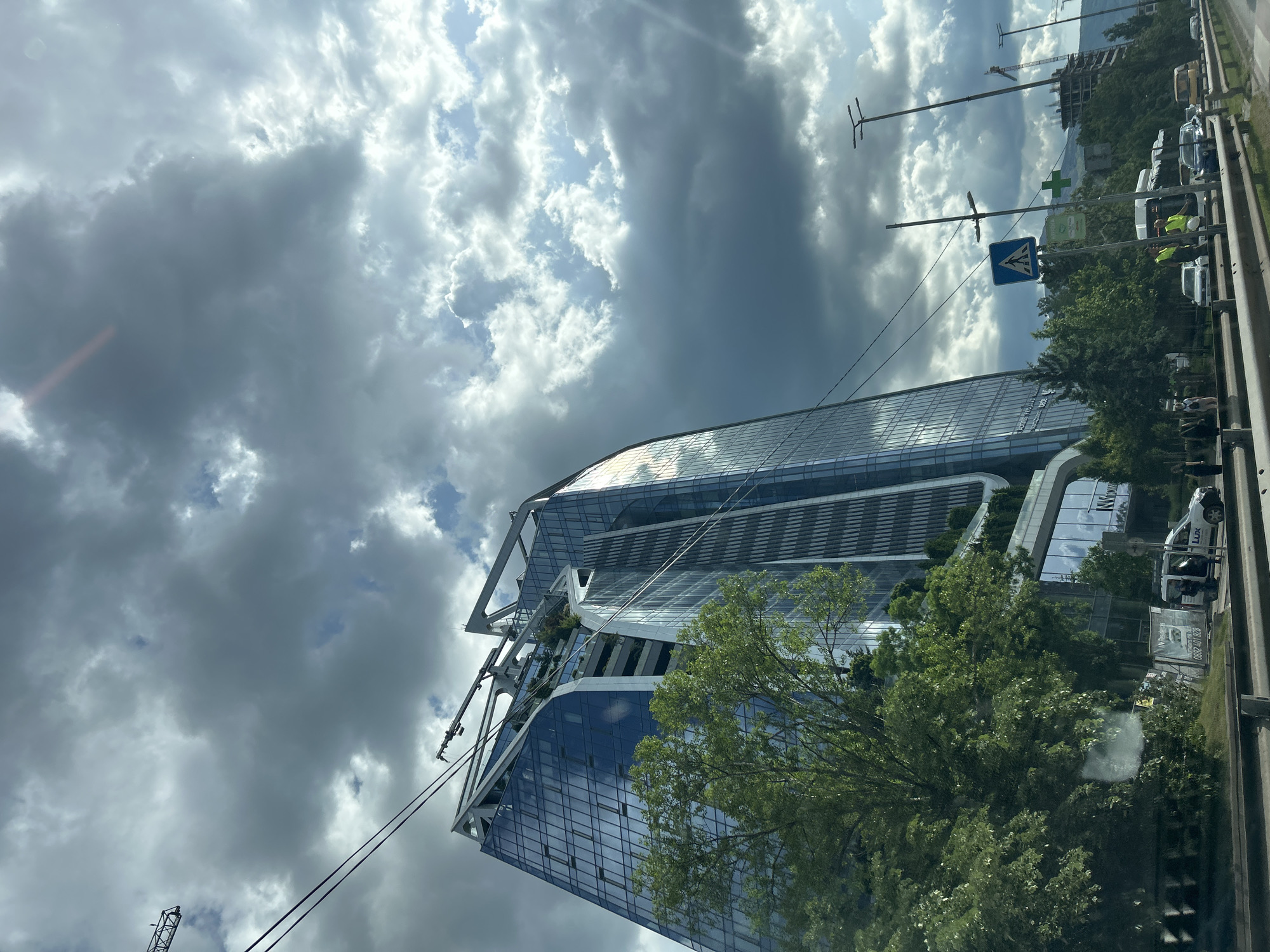 The image depicts a modern glass skyscraper under a sky filled with dark, heavy clouds interspersed with patches of brighter sky. Trees are visible in front of and adjacent to the building, partially obscuring the lower floors. On the right side of the frame, there is a road with cars, a roadside barrier, and street signs. Workers wearing high-visibility vests are seen near the parked vehicles. A building under construction with a crane is visible in the background. The architecture suggests this is the Capital Fort building in Sofia, Bulgaria.
