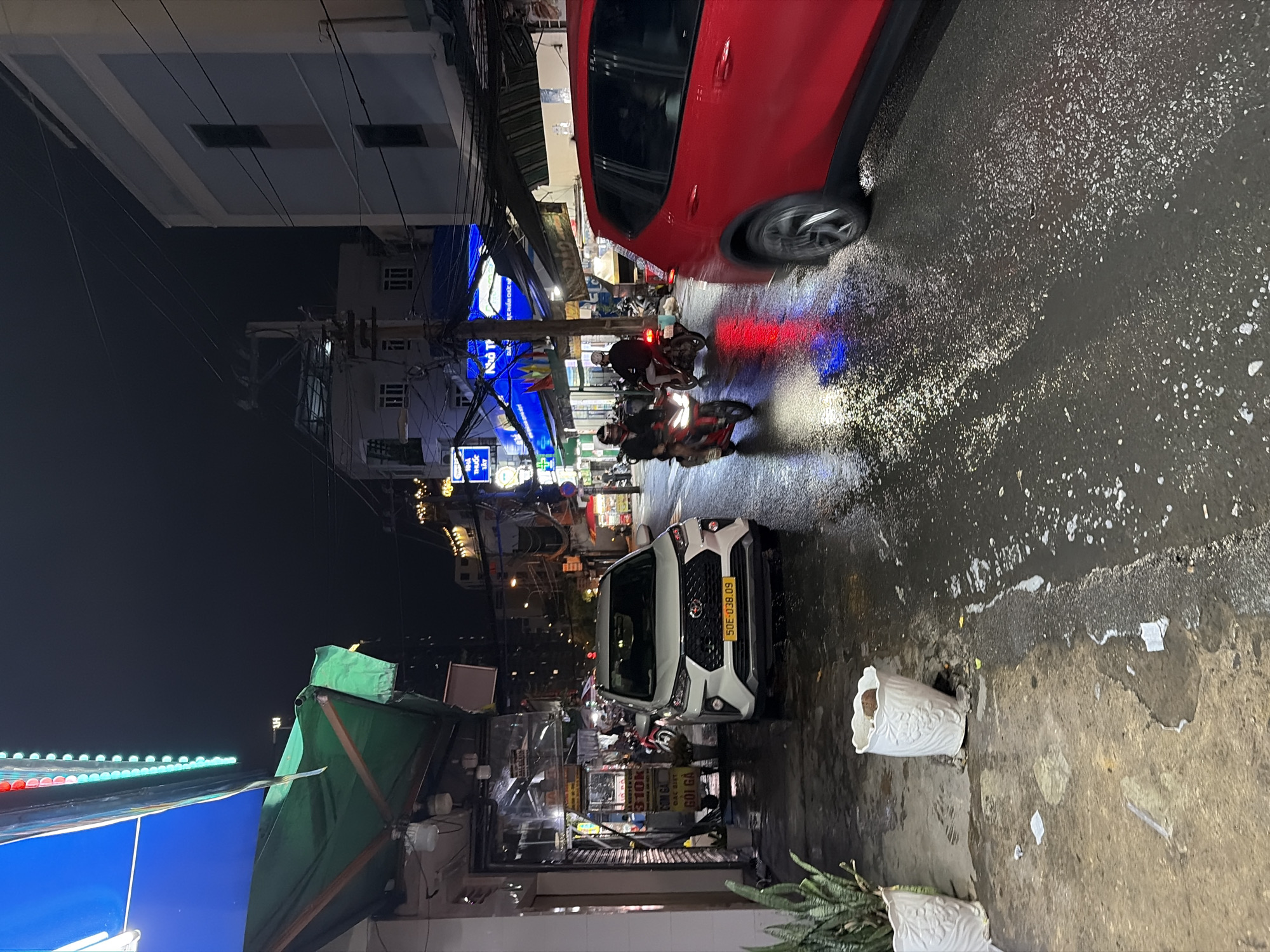 The image depicts a vibrant street scene at night in an urban setting, likely in Thủ Đức, Vietnam, given the visible text. The ground is wet and reflective, suggesting recent rain.
**Subjects:** The foreground features a red car partially visible on the left, and a white SUV (Hyundai Creta or similar) parked on the right, displaying a Vietnamese license plate "50E-038.09". Several motorcycles, ridden by individuals, are navigating the wet street. One rider is clearly visible wearing a helmet and dark clothing. Buildings with multiple stories line both sides of the street, housing various shops and businesses. On the sidewalk, two white decorative planters are visible, one upright and one tilted. Overhead, a dense network of electrical wires crisscrosses the scene.
**Activity or event taking place:** The scene captures typical nighttime street activity. Vehicles are either parked or in motion, indicating active traffic. Shops are open, illuminated by neon signs, suggesting ongoing commerce.
**Notable details:** The time of day is night, characterized by the dark sky and extensive artificial lighting from streetlights, vehicle headlights and taillights, and glowing shop signs. The wet asphalt brilliantly reflects these lights, creating vibrant streaks of red and blue. The architecture and the prevalence of motorcycles are strong indicators of a Southeast Asian city.
**Visible text:** Several signs display Vietnamese text. A prominent blue sign reads "NHÀ THUỐC TÂY" (Pharmacy). Another blue sign partially reads "BIA..." (Beer...). A yellow and white sign near the white SUV shows "310K", "CƠM GÀ", and "GỎI GÀ" (likely referring to 310,000 VND, Chicken Rice, and Chicken Salad, respectively).