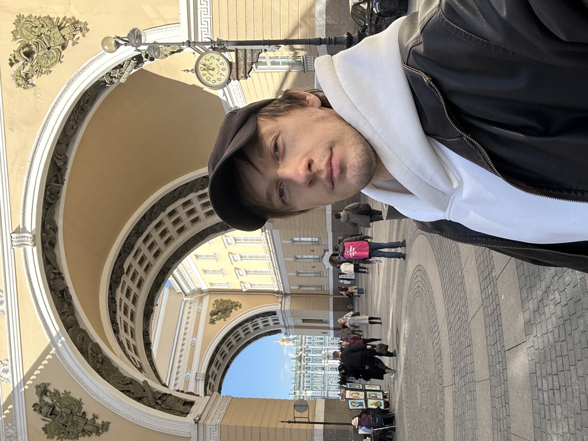 A selfie-style portrait features a young man in the foreground, captured at a slight angle, with his head tilted. He wears a black baseball cap, a white hoodie, and a black leather jacket. His expression is neutral to slightly contemplative.
Behind him, a grand architectural setting unfolds, identified as the Arch of the General Staff Building in Saint Petersburg, Russia. The archway is made of light yellow stone with intricate white and gold decorative elements and carved details. Hanging from the arch structure is a vintage-style street clock with Roman numerals, indicating a time around 10:15 or 10:20.
Through the arch, a bustling public square is visible under a clear blue sky. Numerous people are seen walking and congregating. In the distance, the iconic green and white facade of the Winter Palace, topped with golden domes, can be clearly seen, further confirming the location as Palace Square. The ground is paved with cobblestones and patterned tiles. On the right side of the archway, near the bottom, an outdoor vendor has set up an easel displaying various artworks for sale. The bright sunlight casts distinct shadows, suggesting a clear day, likely late morning or early afternoon.