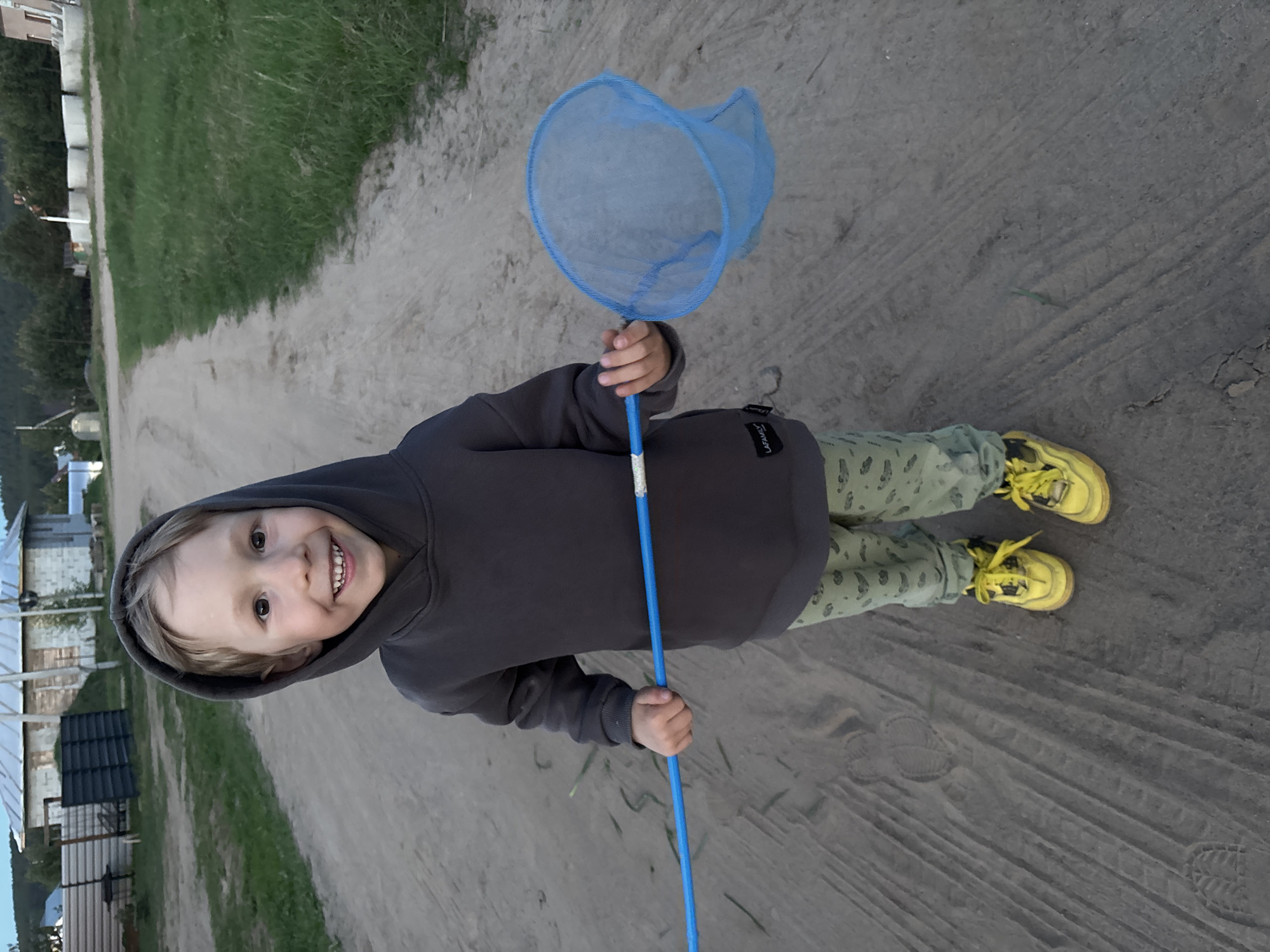 A full-body shot shows a smiling child holding a blue butterfly net while standing on a dirt road. The child wears a gray hoodie, patterned green pants, and yellow sneakers. The dirt road is surrounded by green vegetation and buildings in the distance. The photo was taken in Chaikovsky, Russia.