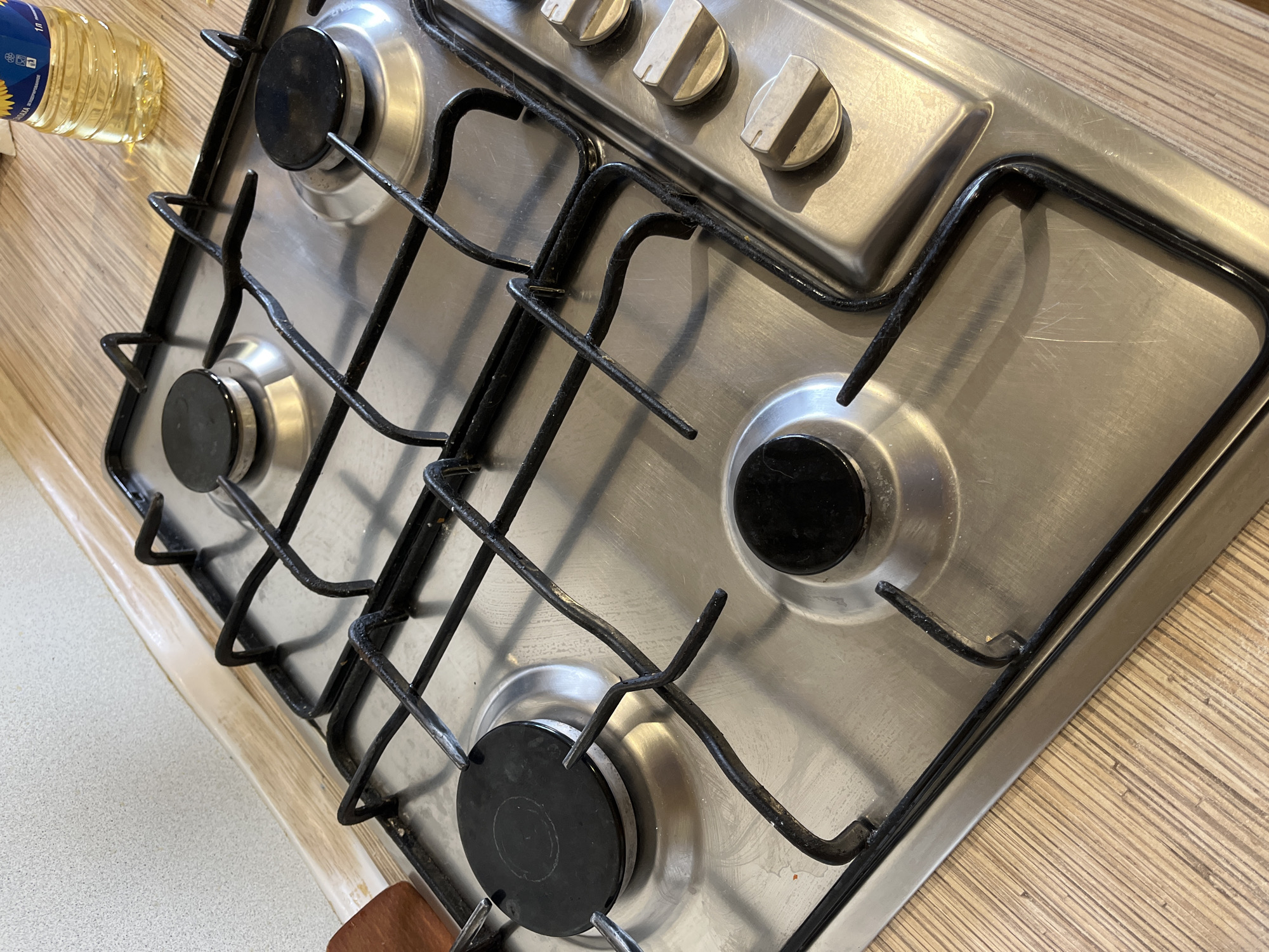 The image displays a close-up, high-angle view of a four-burner gas cooktop integrated into a kitchen counter. The cooktop has a brushed stainless steel surface with black, cast iron grates over each burner and black burner caps. Three light-colored, possibly beige or off-white, control knobs are visible along the top edge of the appliance.
To the upper left of the cooktop, a partially visible clear plastic bottle filled with yellow liquid, identified as sunflower oil, rests on a light wooden countertop. The bottle features a label with a sun logo and Cyrillic text, including "СОЛНЕЧНОЕ" (Solnechnoye, meaning "Sunflower") and "1л" (1 liter), indicating a Russian-speaking context, consistent with Chaikovsky, Russia. The wooden countertop extends around the back and left side of the stove, while a different, lighter speckled countertop is visible to the right.
The stainless steel surface of the cooktop shows signs of use, including minor scratches and scuff marks. Some light residue is also visible on the black grates. No cooking activity is taking place; the stove is off. The setting is indoors, likely a kitchen. No people are visible.