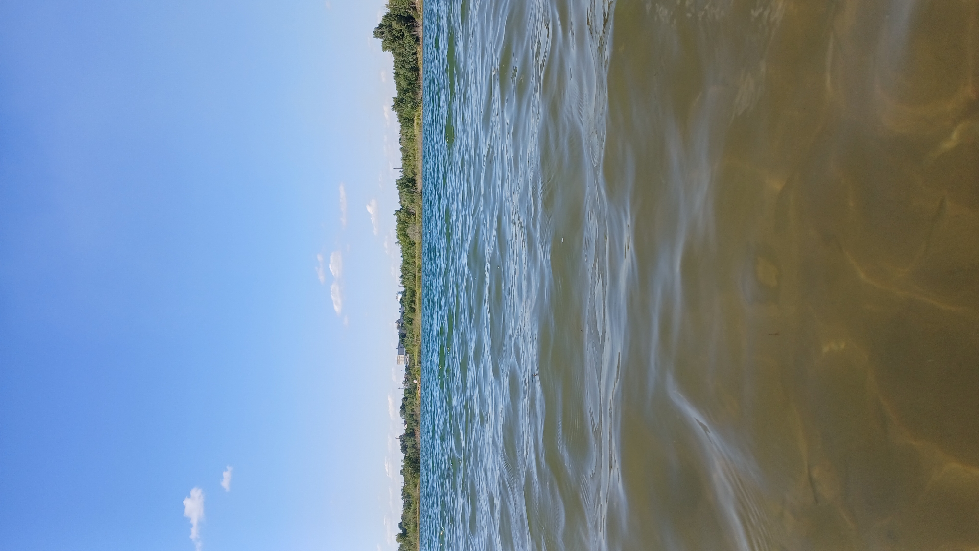 The image depicts a body of water with gentle waves, likely a lake or a large pond, under a clear blue sky with a few scattered white clouds. The water in the foreground is relatively shallow, revealing the sandy bottom with sunlight creating dappled patterns on it. As the view extends to the horizon, the water appears deeper and takes on a turquoise hue. Along the distant shoreline, a line of trees and vegetation is visible, with some buildings appearing further inland. The scene suggests a natural outdoor environment, potentially a recreational area, within or near the city of Jelaevo, Kazakhstan. There are no people or specific actions depicted in the image.