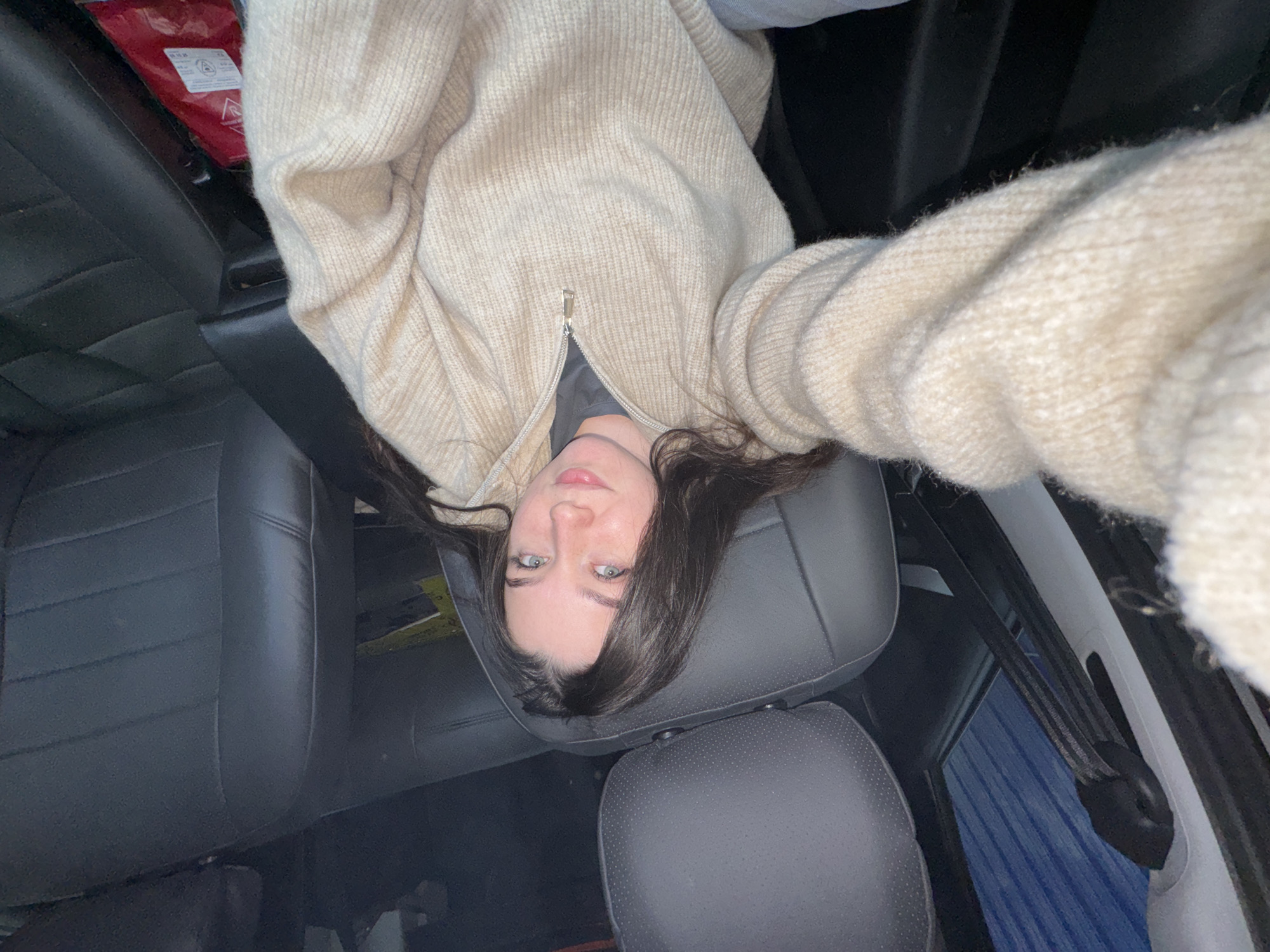 A young woman is depicted in a selfie taken inside a car, likely at night or in a dimly lit environment, possibly in Perm, Russia. She is looking directly at the camera with a neutral, calm expression, showcasing her blue-green eyes. Her long, dark hair frames her face as she appears to be lying down or leaning back, resting her head on a black, perforated car seat headrest. She is wearing a cream-colored, ribbed, zippered cardigan over a dark top.
The car's interior features dark grey or black leather seats. A black seatbelt is visible on the right side of the frame. In the upper left background, behind the woman's head, a red fire blanket is clearly visible. It is folded and positioned, featuring a white label with black text. Notable text on the label includes "EN 1869," which is a European standard for fire blankets, along with warning symbols such as a flame graphic and instructional arrows. The overall lighting is soft and localized, highlighting the woman's face against the darker vehicle interior. The image captures a casual, intimate moment within the confined space of a vehicle.