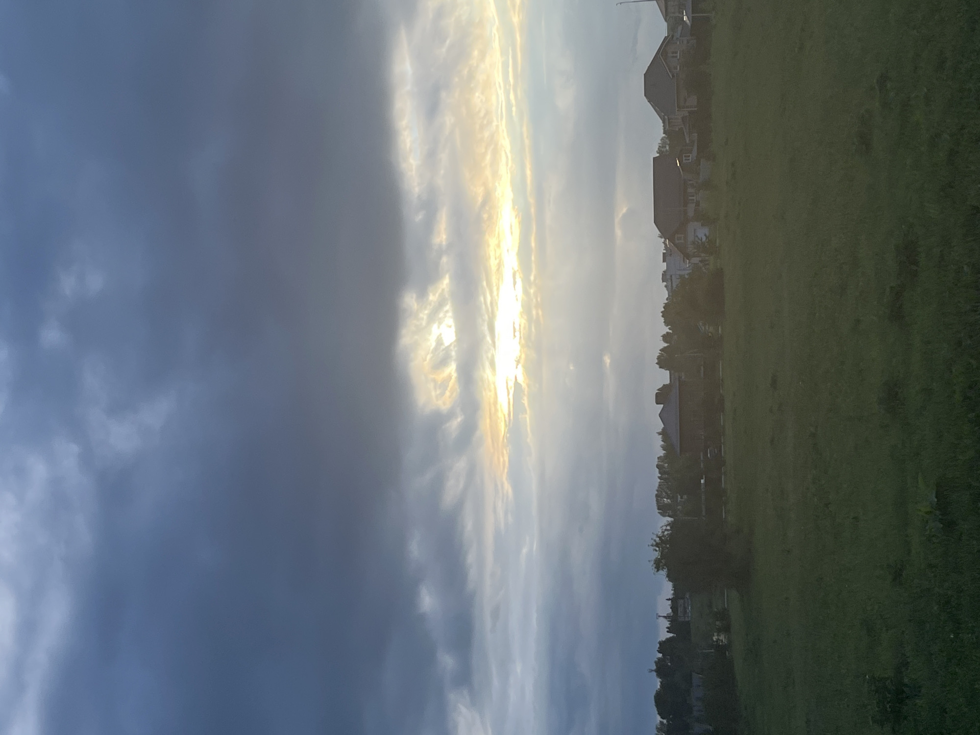 This is a vertical, wide-angle photograph of a suburban landscape at sunset. The sky dominates the left half of the frame, filled with dramatic clouds ranging from dark, ominous grey on the far left to lighter, wispy formations illuminated by the setting sun on the right. The sun itself is a bright, almost blinding glow peaking through the clouds, casting a warm, golden light that reflects off the cloud edges.
To the right of the sky, a line of houses with peaked roofs is visible, nestled amongst trees. The houses appear to be typical suburban dwellings, and their architecture suggests a European or North American setting, consistent with the Kamskiye Polyani location. The trees are leafy and green, indicating a summer or late spring season.
The right half of the image is a large, open field of green grass. The grass is somewhat uneven and appears to be a park or undeveloped lot. The overall impression is one of a peaceful, end-of-day scene in a quiet residential area. The lighting suggests it is late evening, around sunset, with a mix of cloudy and clear skies. There is no visible text in the image.