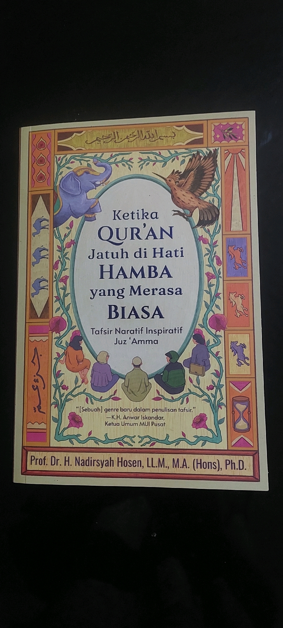 A close-up, overhead shot shows the cover of a book titled "Ketika QUR'AN Jatuh di Hati HAMBA yang Merasa BIASA". The cover features an illustration with an elephant and a bird. It also includes the subtitle "Tafsir Naratif Inspiratif Juz 'Amma" and a quote attributed to K.H. Anwar Iskandar, Ketua Umum MUI Pusat. The author's name, "Prof. Dr. H. Nadirsyah Hosen, LL.M., M.A. (Hons), Ph.D.", is printed at the bottom. The book cover is laid flat on a black surface, likely a table or floor. There is no indication of the city of Lumajang, Indonesia, or any specific location within it.