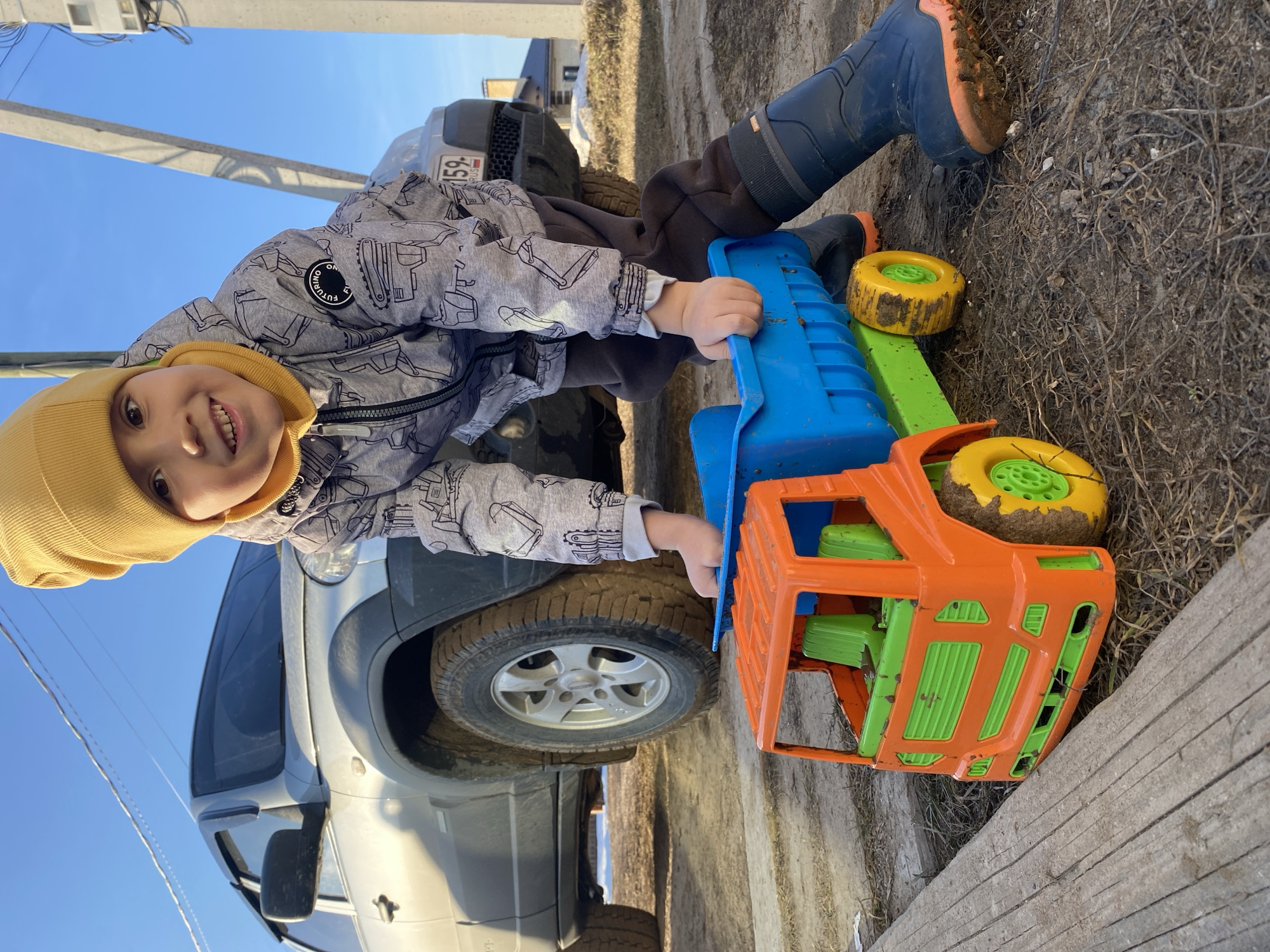 A young boy wearing a yellow beanie, a gray jacket with construction vehicle designs, brown pants, and blue boots is playing with a large, colorful toy truck. The truck is primarily orange, blue, and green, with yellow wheels that are muddy. The boy is smiling at the camera. A gray SUV is visible behind the boy and the toy truck. The license plate of the SUV reads "159" and appears to be registered in Russia. The location seems to be a partially muddy area with some dry grass and what looks like a wooden board in the foreground. Power lines are visible in the background against a blue sky. The city of Chaikovsky, Russia, is given as the location.