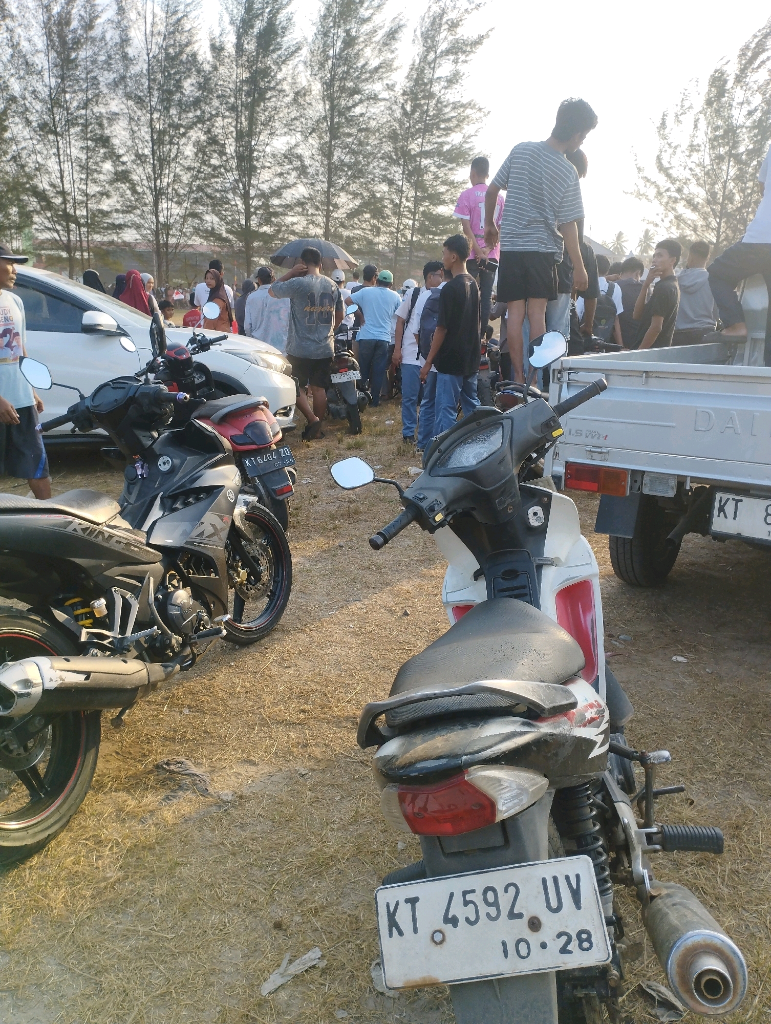The image depicts an outdoor scene, possibly a parking lot or an open field, with a group of people and several motorcycles. In the foreground, a white and red motorcycle is parked facing the viewer, with its license plate clearly visible as "KT 4592 UV 10.28". To the left, a black and red motorcycle is also parked.
A crowd of people, predominantly young men, are gathered in the background. Some are standing, while others appear to be sitting or leaning on a silver truck with "DAIHATSU" branding. The people are dressed casually in t-shirts and shorts. The environment is characterized by dry, sparse grass and several tall trees with needle-like foliage in the background, suggesting a warm climate. The lighting indicates it is daytime, possibly late afternoon, with a bright sky. The overall impression is of a casual gathering or event.