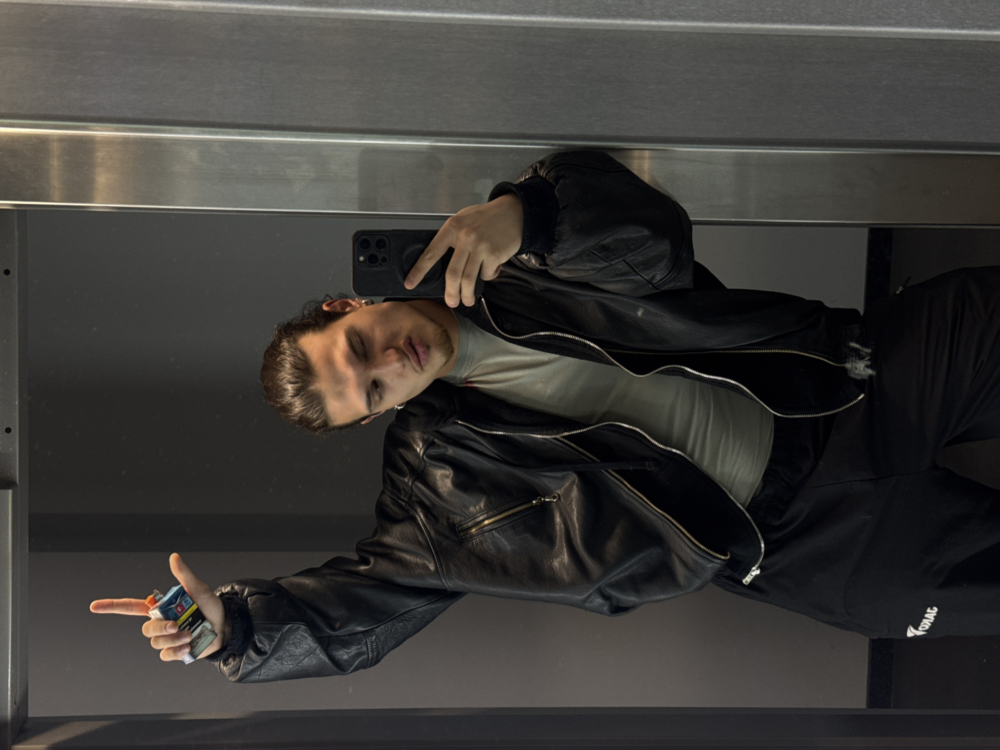 A young man is featured in a mirror selfie taken indoors in Sofia, Bulgaria. He has dark hair and stubble on his chin, with an earring visible in his left ear. He is dressed in a black leather jacket over a light-colored t-shirt and black trousers that display the word "ZODIAC" in white text on the right leg.
In his right hand, he holds a black smartphone with multiple camera lenses, using it to capture the reflection. His left hand holds a small, colorful rectangular object, possibly a pack of cigarettes or a vaping device, while his index finger points outwards. The setting appears to be a modern indoor space, likely a hallway, elevator, or restroom, indicated by the large reflective surface and metallic horizontal panels at the top and bottom of the frame. The lighting is artificial and somewhat dim. The man maintains a neutral to slightly serious expression, with his lips slightly parted, as he poses for the camera.